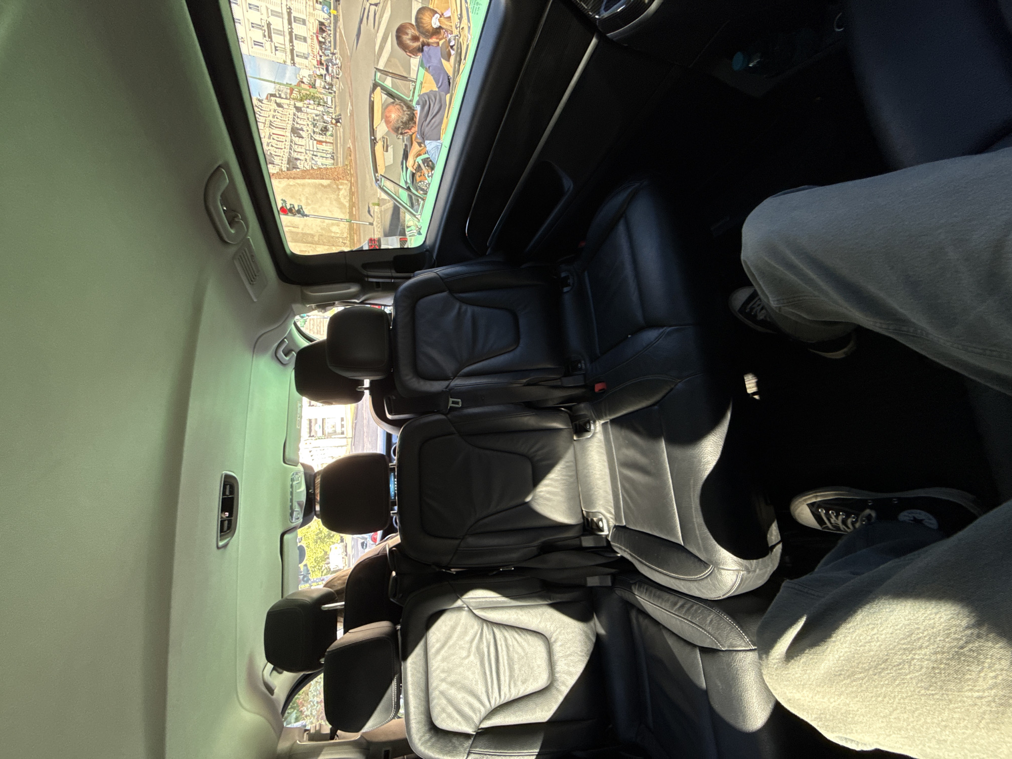 The image shows the interior of a car, specifically the back seats, from a slightly tilted perspective. The seats are black leather and arranged in three rows. Two people are sitting in the back seats of the car, wearing jeans and shoes. The car appears to have a sunroof, through which a street scene in Milan, Italy, is visible. There is a green car, two people with blonde hair, and an adult in the neighboring lane. The architecture visible through the sunroof suggests an urban environment. A traffic light can also be seen.