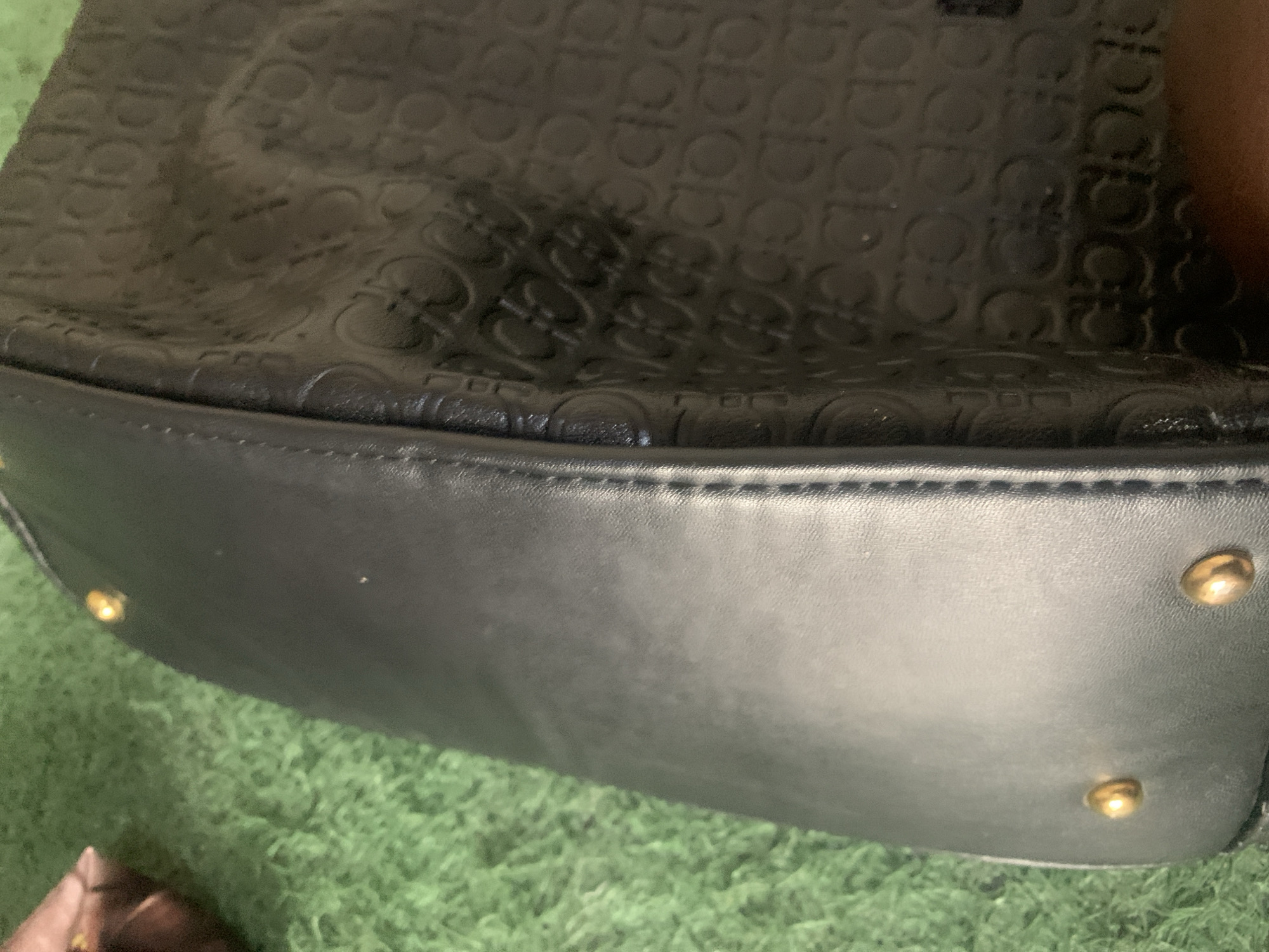 The image shows a close-up of a black leather handbag with an embossed pattern of interlocking "G" logos. The handbag has gold-toned studs along its edge. The texture of the leather and the pattern are clearly visible. The handbag is resting on a green, textured surface that appears to be artificial grass or a similar material. A blurred object, possibly a hand or another item, is visible in the lower right corner of the image. The lighting appears to be from above, casting subtle shadows that highlight the embossed details. There is no indication of the location being Jauro Yinu, Nigeria; the description is solely based on the visual elements present in the image.