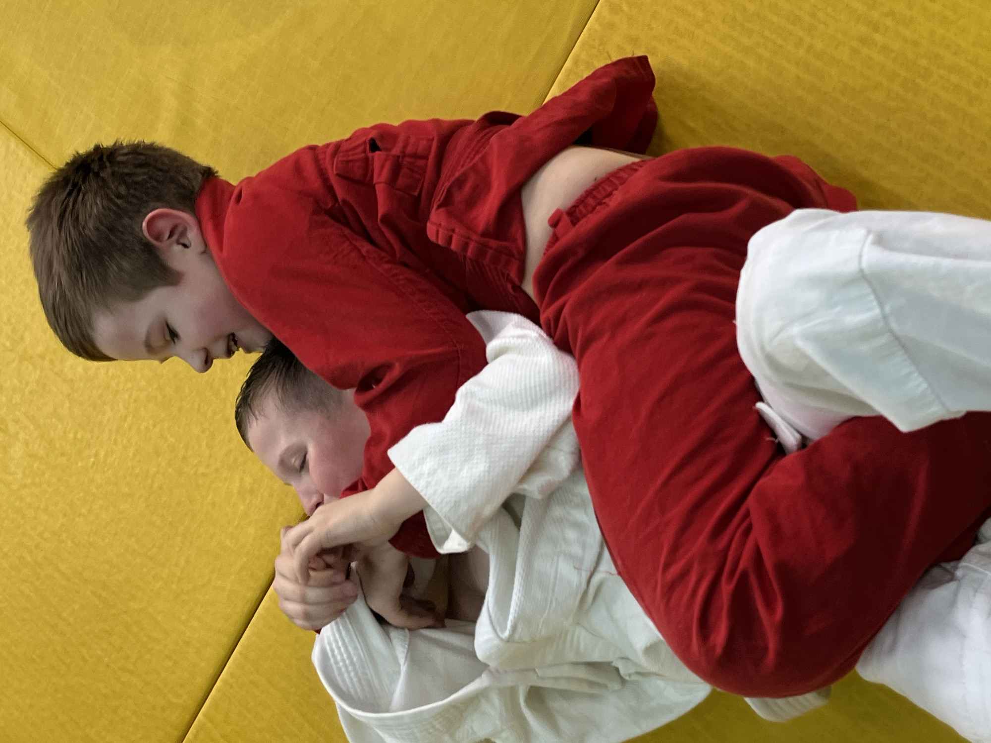 Two young boys are actively engaged in a grappling or sparring session on a bright yellow padded mat, indicative of a martial arts training environment such as a dojo or sports hall in Krasnoyarsk, Russia. One boy, wearing a red martial arts uniform (gi), is positioned on top of the other boy, who is dressed in a white gi. The boy in the red gi is looking downwards and to his left, with a focused expression, possibly a slight smile, suggesting concentration and effort. His left arm wraps around the torso of the boy in white, and his hands are gripping the white uniform, indicating a hold or technique being applied. The boy in the white gi is partially obscured beneath, appearing to be in a defensive or counter-grappling position, with his hands also engaged in gripping the red gi. The lighting suggests an indoor setting, likely during the daytime. The image captures a dynamic moment of physical education or martial arts practice. No visible text is present in the media.
