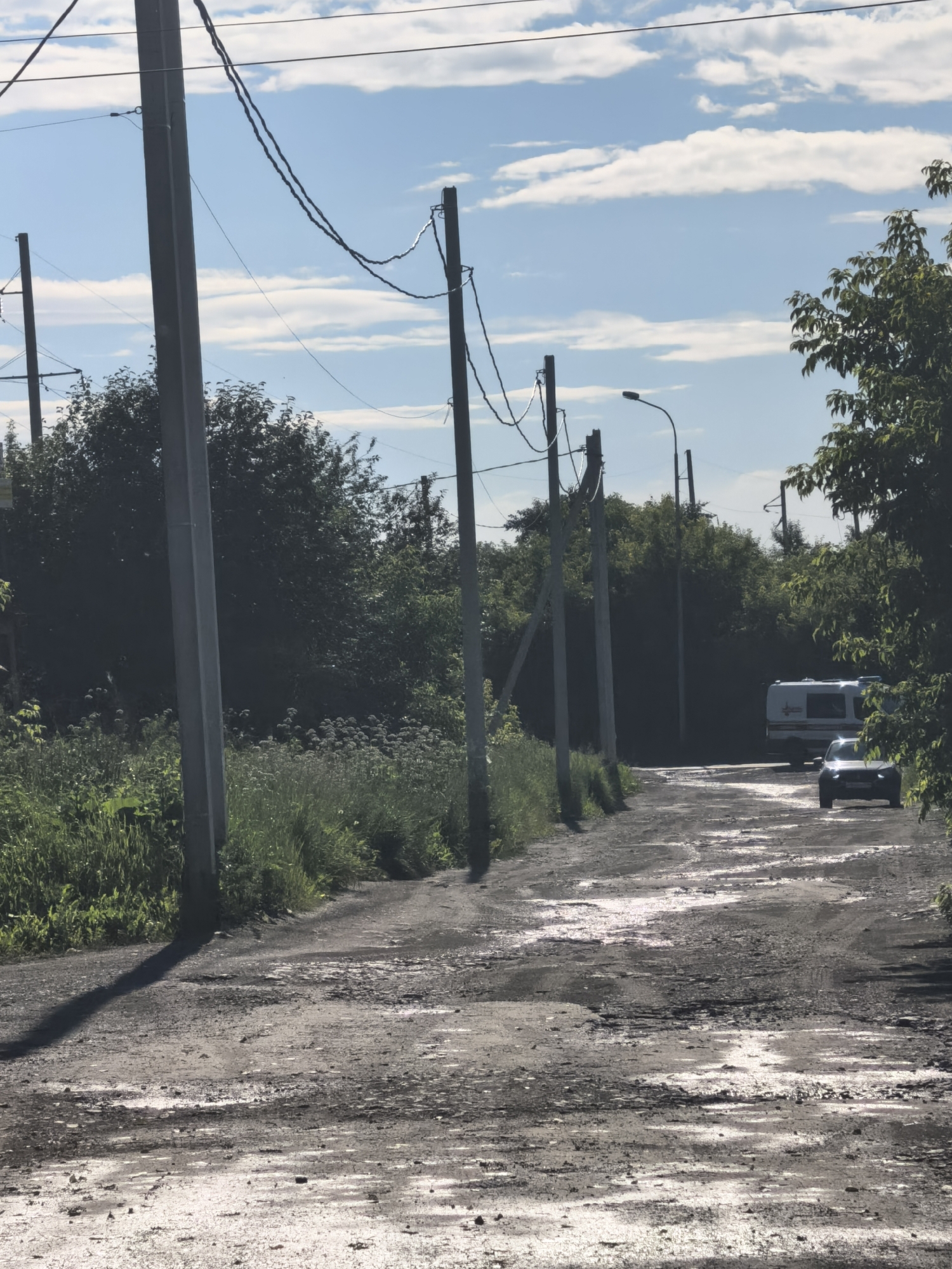 The image depicts a rural road on a sunny day with scattered clouds. The road is unpaved and appears to be in poor condition, with numerous potholes and puddles reflecting the sky. Along the side of the road, tall green grass and bushes are visible, interspersed with power poles and overhead wires. Further down the road, two vehicles can be seen: a white recreational vehicle and a dark-colored car. The presence of power poles and the unpaved road suggest a somewhat undeveloped or rural setting. The sunlight indicates it is daytime, and the clear sky with some clouds suggests fair weather. The scene evokes a sense of a journey or transit through a natural, albeit somewhat neglected, environment. There is no visible text in the image.