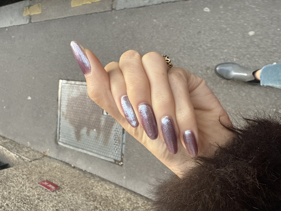 A close-up, high-angle view focuses on an individual's left hand, extended forward on an urban street in Paris, France. The hand features five almond-shaped fingernails, each painted with an iridescent purple-mauve polish that displays a prominent light blue-silver shimmer. A gold-toned ring with an intricate, possibly floral or geometric, openwork design is present on the ring finger. The individual's wrist is obscured by a dark brown, shaggy garment, indicative of fur or faux-fur. At the bottom right, the top of a grey boot and a section of light blue denim jeans are visible, indicating the individual is walking. The background consists of a grey asphalt sidewalk containing a square metal utility access panel. To the left, a textured, light-colored concrete or stone wall runs parallel to the sidewalk, bearing a small, rectangular red sign with unidentifiable white text. Beyond the sidewalk, a grey asphalt street with a faint yellow line is discernible.