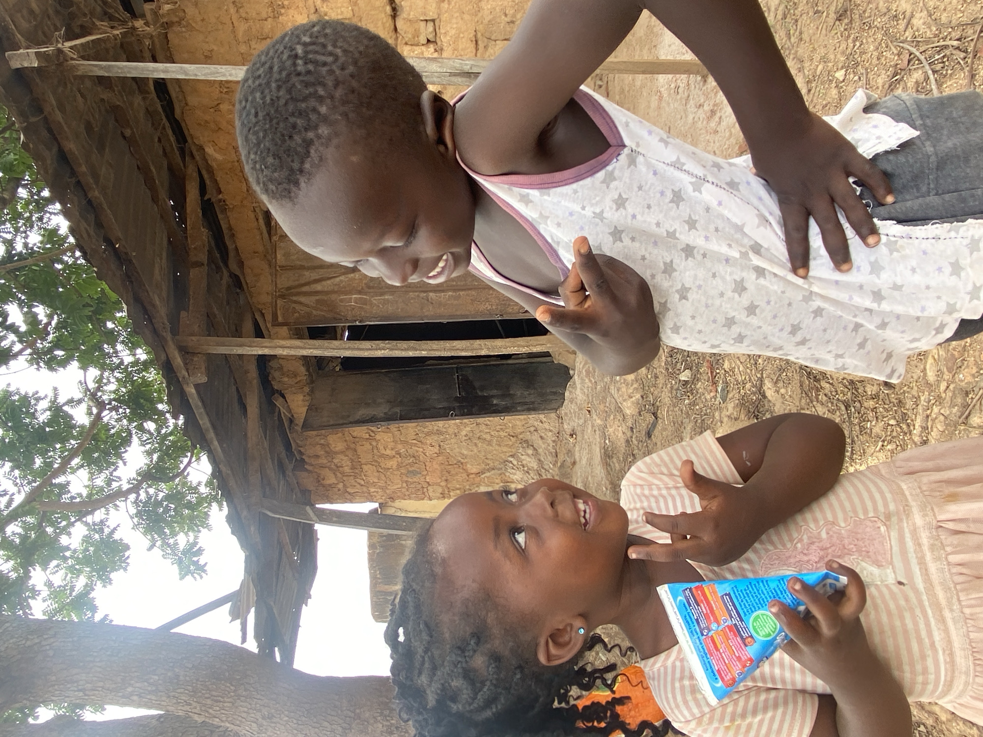 In Kuru Dungus, Nigeria, two young girls are facing each other outside a simple structure. The girl on the bottom is holding a tetra pack-style container. They are both smiling. The building behind them appears to be constructed with wood. A tree with green leaves partially obscures the upper left portion of the frame. The ground appears to be dirt.