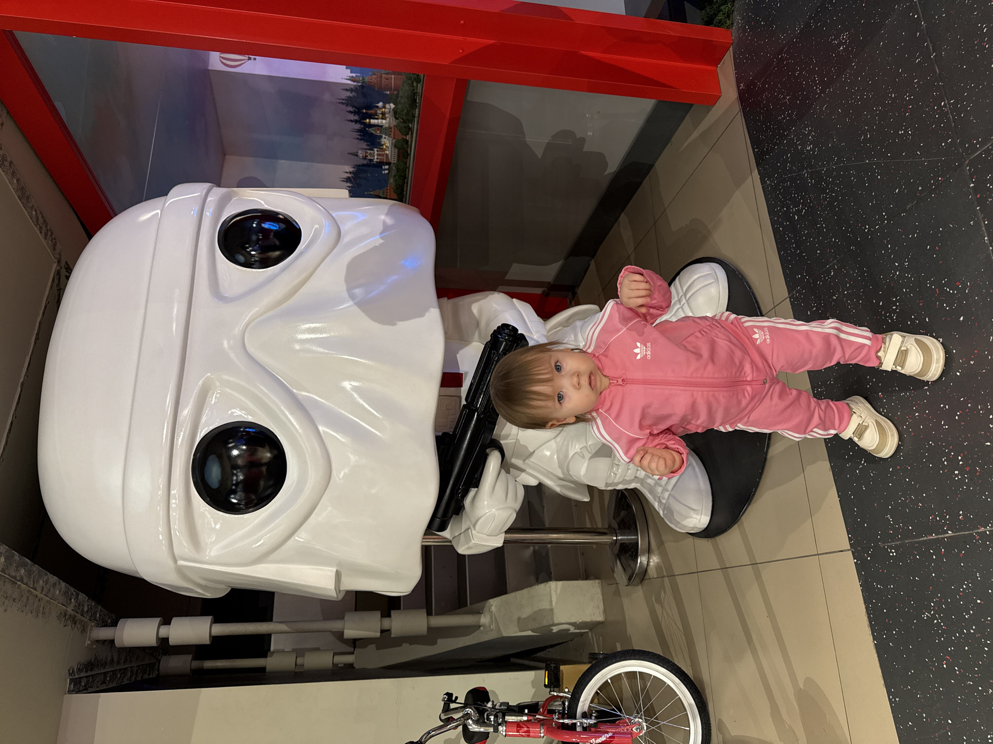 A young child is laying on the lap of a life-sized Stormtrooper statue. The child is wearing a pink Adidas track suit and tan shoes. The Stormtrooper is holding a black gun. To the left of the Stormtrooper is a red children's bicycle. The location is in the city of Unknown, Unknown.