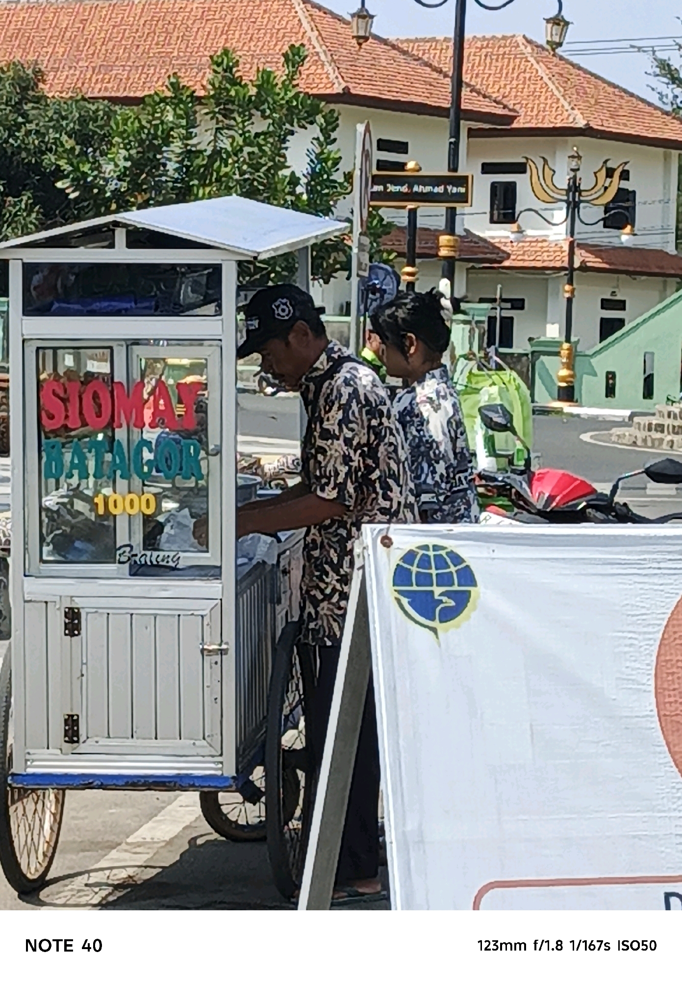 A mobile food cart, displaying signs for "SIOMAY BATACOR" and a price of "1000", is parked outdoors on a street in Desa Tambakreja, Indonesia. A man wearing a patterned shirt and a black baseball cap is operating the cart. To his right, a young woman in a patterned shirt stands by. In the background, a building with an orange tiled roof and a street sign reading "Jl. Jend. Ahmad Yani" are visible. A motorcycle with a red seat is parked behind the food cart. A white banner with a blue globe logo is positioned in front of the cart. The scene is illuminated by bright daylight.