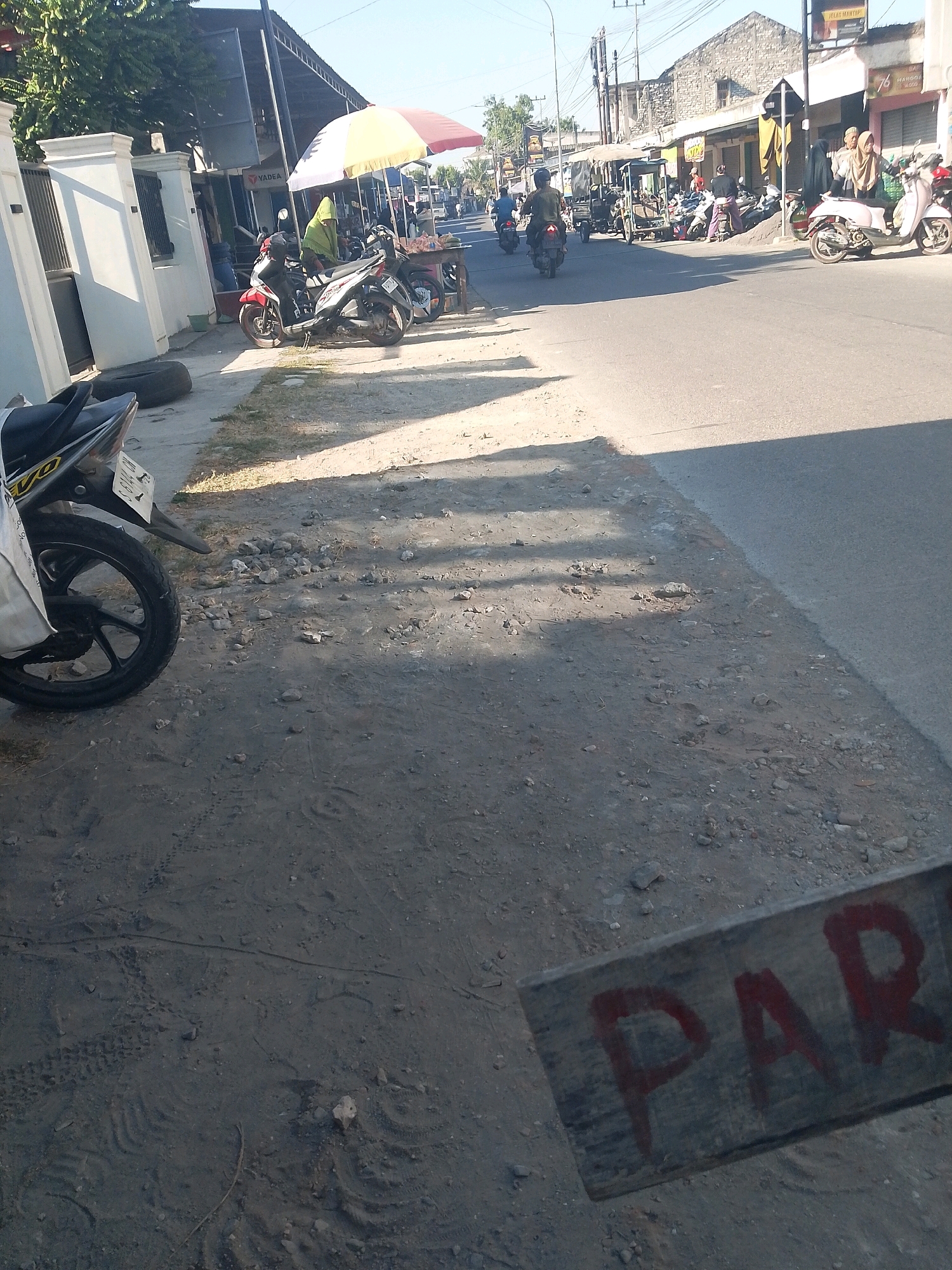 The image captures a street scene in Jampet, Indonesia, during daylight. The foreground shows a dusty, uneven ground surface with scattered stones and tire tracks. A weathered wooden plank with the partially visible red painted word "PARK" lies diagonally across the lower right portion of the frame. To the left, the rear wheel and fender of a black motorcycle are visible, with a white bag hanging from it.
Further into the scene, a row of shops line the street on the left side, with a white fence and gate in front of one establishment. A red and yellow umbrella is set up near the shops, shading a market stall where a person wearing a green headscarf is interacting with a vendor. Several motorcycles are parked along the roadside.
The street itself is paved and shows signs of moderate traffic. In the background, more buildings and parked vehicles can be seen, suggesting a bustling commercial area. The sky is clear and bright, indicating sunny weather. The overall impression is of a typical street environment in a town or city in Indonesia.
