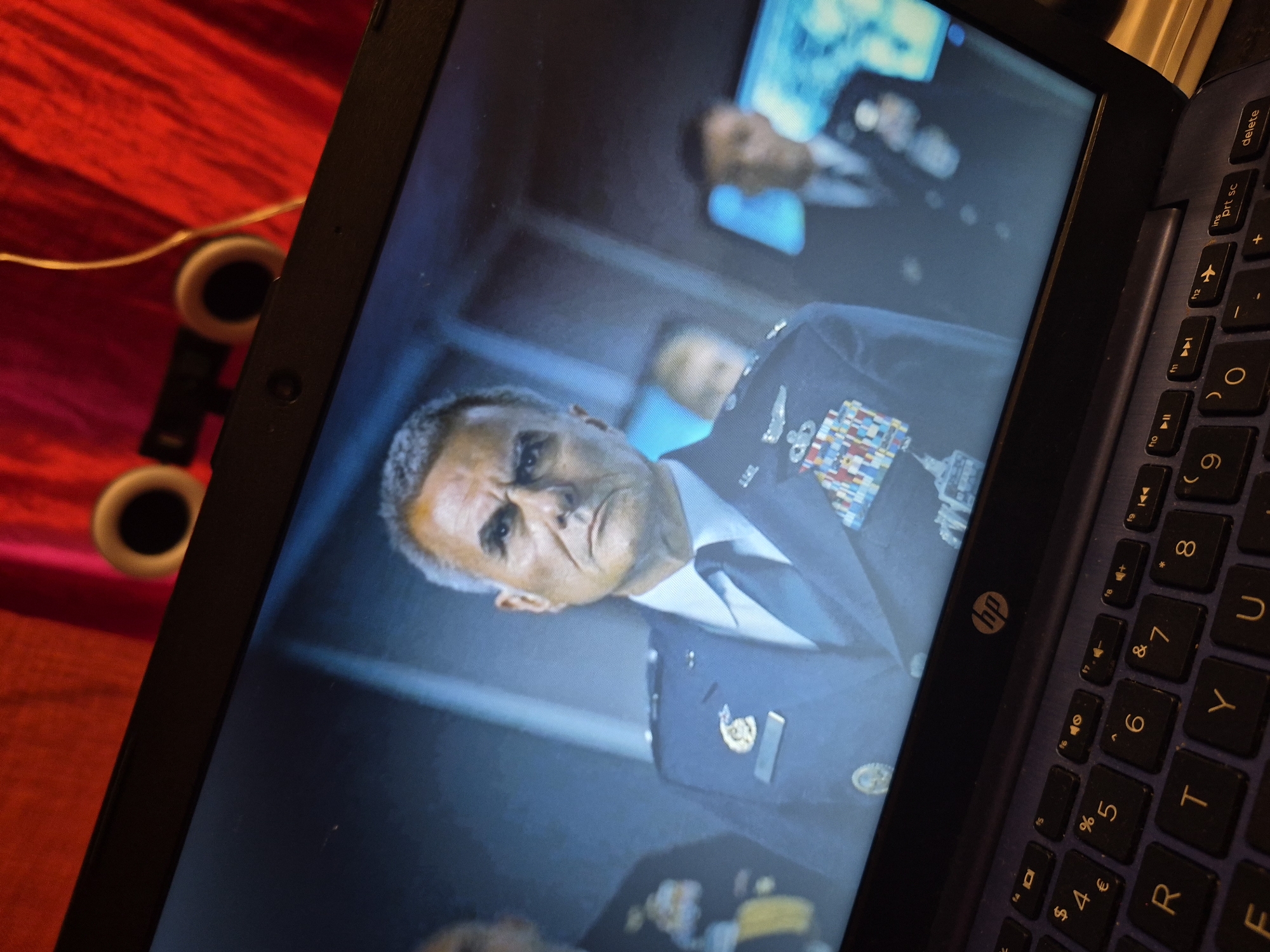 A close-up, high-angle shot shows a laptop with a digital image displayed on its screen. The screen shows a man in a military uniform, wearing multiple medals and a tie, looking towards the viewer with a stern expression. Behind him, other individuals in uniform are blurred and less distinct. The laptop's keyboard is visible in the foreground, with various keys including letters, numbers, and function keys. The laptop itself appears to be dark blue. In the background, out of focus, is a red, textured surface and two earbud-like objects with a thin wire connecting them. There is no indication of the location being in the city of Sandwell, United Kingdom.