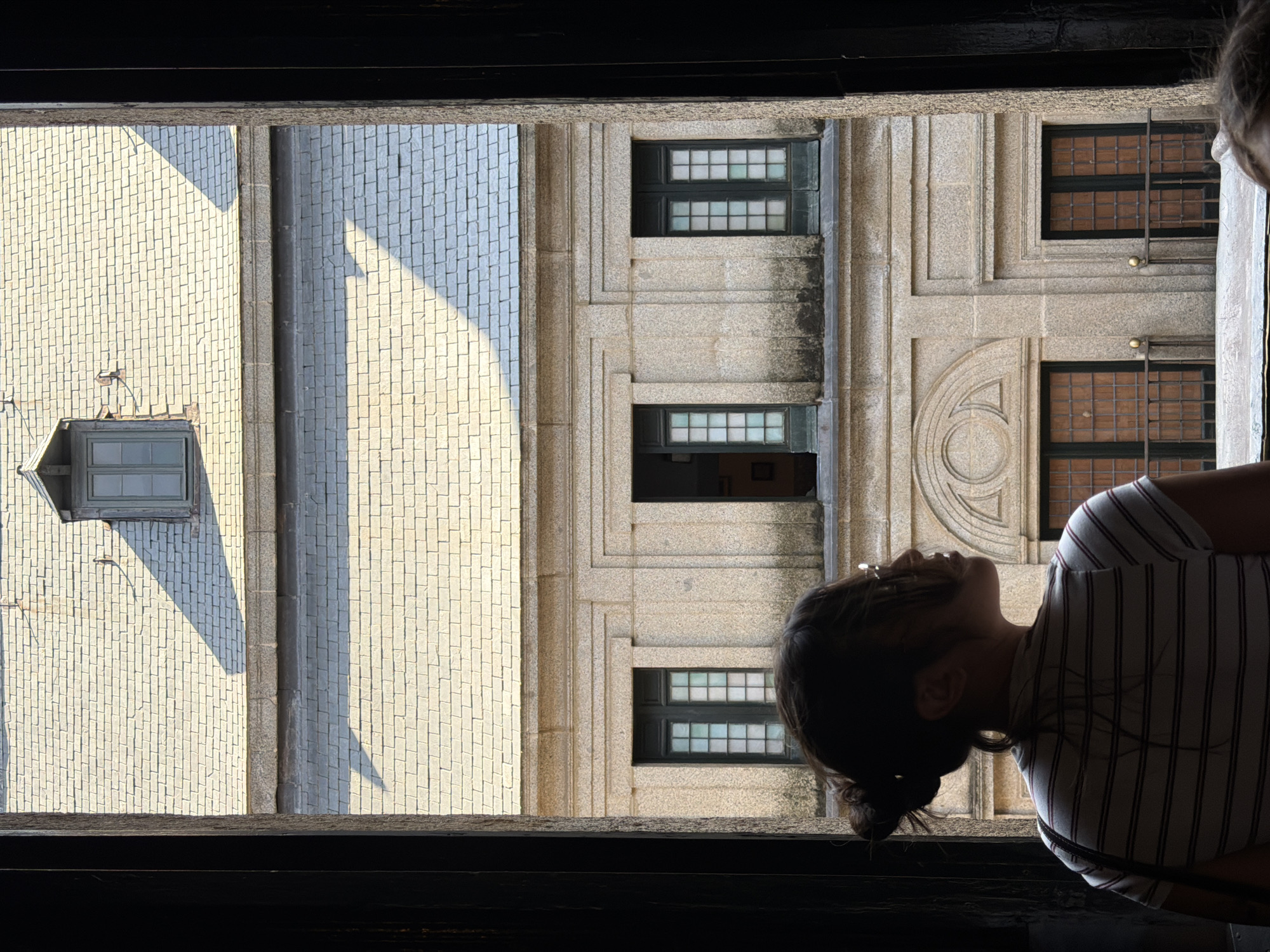 This is a photograph taken from inside a dark doorway, looking out at a stone building with a grey tiled roof and a dormer window. In the foreground, silhouetted against the light, is the back of a person with their hair tied up in a bun. They are wearing a striped t-shirt and are looking to the right. The building facade has multiple windows, some with dark frames and gridded panes, and others with closed wooden shutters. The sunlight casts sharp shadows on the roof and facade, indicating a bright day. The architecture suggests a historical or institutional building. The scene is located in San Lorenzo De El Escorial, Spain, likely within the Royal Site of San Lorenzo de El Escorial, a UNESCO World Heritage site that includes a palace, basilica, monastery, and museum.