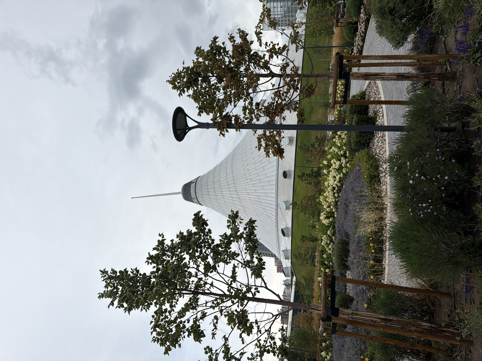 The image shows a view of the Khan Shatyr Entertainment Center in Astana, Kazakhstan. The distinctive tent-shaped building is visible in the background, with its white, translucent roof reaching towards a cloudy sky. In the foreground, a well-maintained park area features several young trees, some with wooden supports, a lamppost, and a variety of flowering plants, including lavender and white hydrangeas. A paved pathway winds through the greenery. The scene appears to be a public space, designed for leisure and enjoyment, with no people present. The overall atmosphere is serene and the lighting suggests an overcast day.