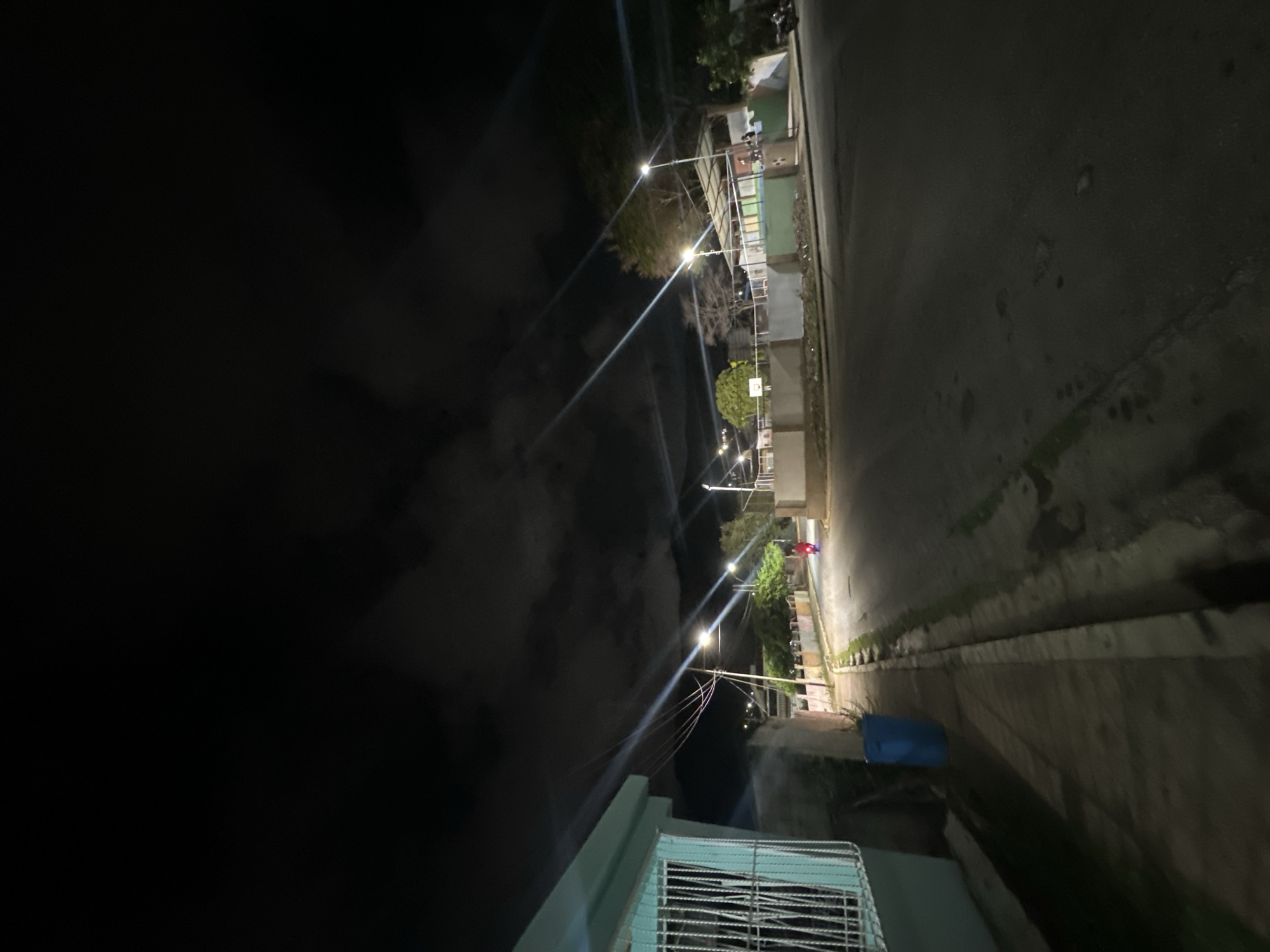 An outdoor, nighttime street scene in El Maco, Venezuela. A turquoise building with a white barred window is on the left. A blue trash can is next to the building. A street runs diagonally into the background, lined with houses and streetlights. A person on a red motorcycle is traveling down the street. The sky is dark with clouds and illuminated by light beams from the streetlights.