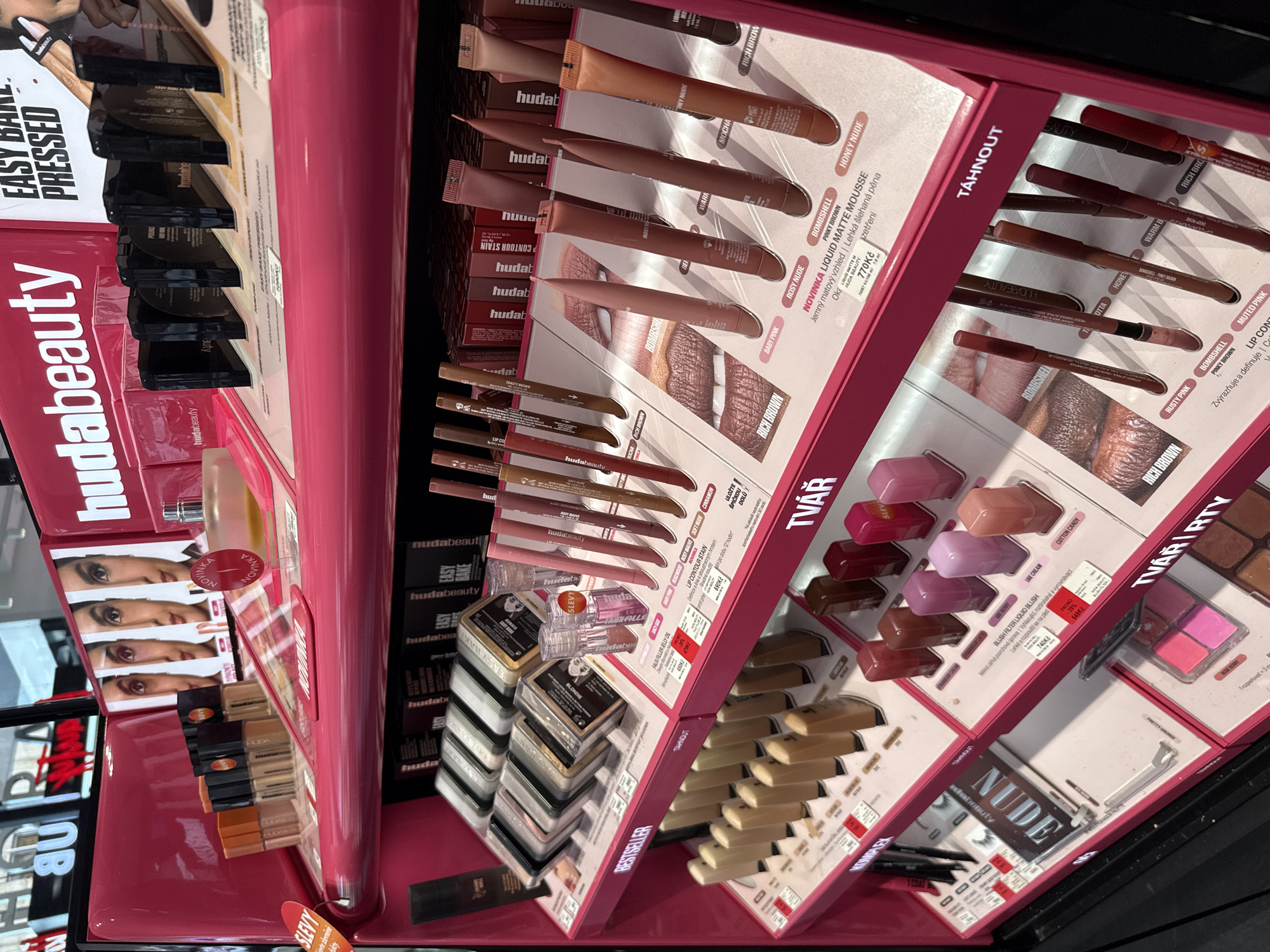 The image depicts a display of Huda Beauty cosmetics in a retail setting. The products are arranged on pink shelves, with a focus on lip products and foundations. Several shades of liquid lipsticks and lip liners are prominently displayed, along with compact powders and other makeup items. The display includes promotional material with images of models and product names. The setting appears to be a department store or a dedicated beauty store. There are no people visible in the image. The lighting is bright, likely from overhead store lights, highlighting the colors and packaging of the products. The overall impression is one of a well-organized and attractive retail display.