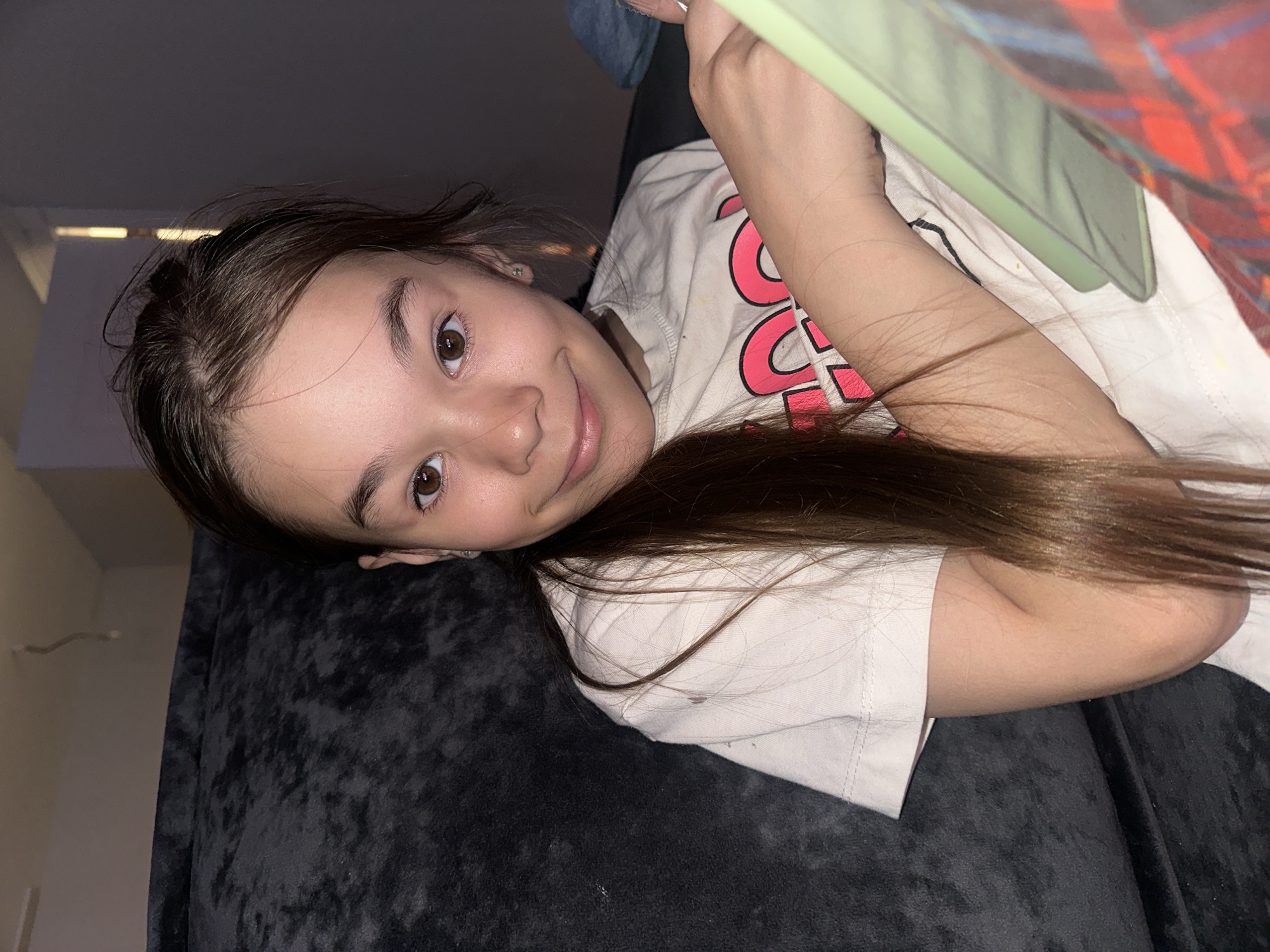 A young girl with brown hair lies on a dark gray textured surface, possibly a couch or bed. She is tilted at an angle towards the camera, smiling slightly. She is wearing a light-colored t-shirt with pink and red lettering. In her hands, she holds a tablet with a light green case. The background includes a window and a wall, with a light fixture visible on the wall. There is no indication that this image was taken in Kharnavy, Russia.