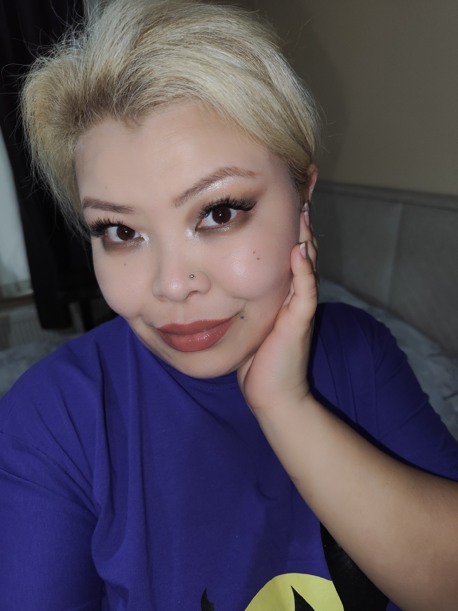 A close-up selfie shows a woman with short, blonde hair, fair skin, and brown eyes. She is wearing a purple t-shirt. Her left hand is gently touching her cheek. She has a nose piercing and a mole on her face. Her makeup includes eyeshadow, eyeliner, and a light brown lipstick. The background appears to be an interior setting, possibly a bedroom. The location in Sarıyer, Turkey, cannot be determined from the image.