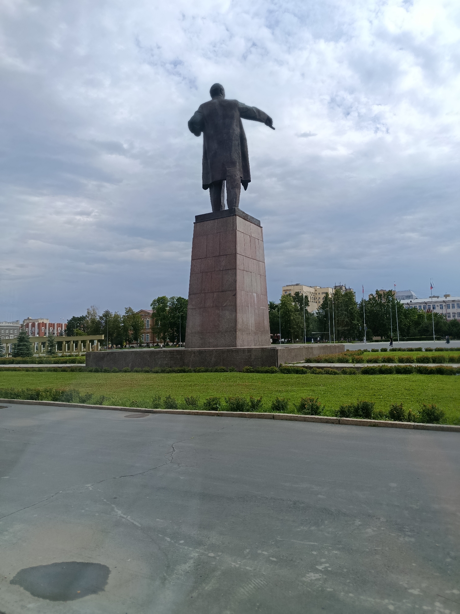 A tall, imposing bronze statue of Vladimir Lenin stands on a large, multi-tiered stone pedestal. Lenin is depicted from behind, facing away from the viewer, with his right arm extended forward and his left arm bent at the elbow, holding what appears to be a hat. The statue and pedestal dominate the foreground and mid-ground of the image.
Behind the monument, a well-maintained green lawn stretches out, bordered by low bushes. Further in the background, several buildings are visible, including a large, light-colored building with multiple windows on the right, and a row of older, ornate buildings with red brick accents and classical architectural features on the left. Trees are interspersed between the buildings and along the horizon.
The sky is overcast with heavy, grey clouds, suggesting a potentially cool or damp day. The lighting is diffused, indicating it is likely daytime, but not particularly sunny. The scene appears to be a public square or park in Saratov, Russia, given the architectural styles of the buildings and the presence of a prominent Lenin statue, a common feature in many Russian cities. No specific activity or event is clearly depicted; the scene appears static, focusing on the monument and its surroundings. There is no visible text in the image.