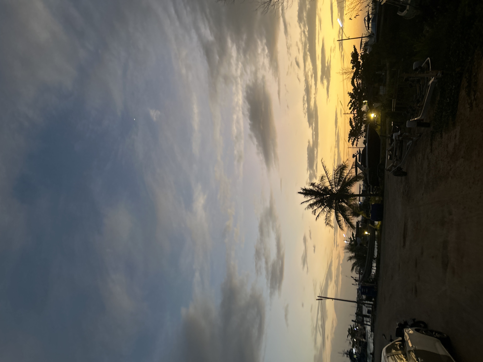 This is a photograph of a harbor area in Ko Pha-ngan District, Thailand, during dusk. The sky is a gradient of blue and orange hues, with scattered clouds. In the foreground, there is a dirt road with a white and black car on the left. Various boats and boat trailers are visible throughout the scene, some on land and others in the water. A palm tree stands prominently in the middle ground. The overall impression is a serene, twilight ambiance in a coastal location.