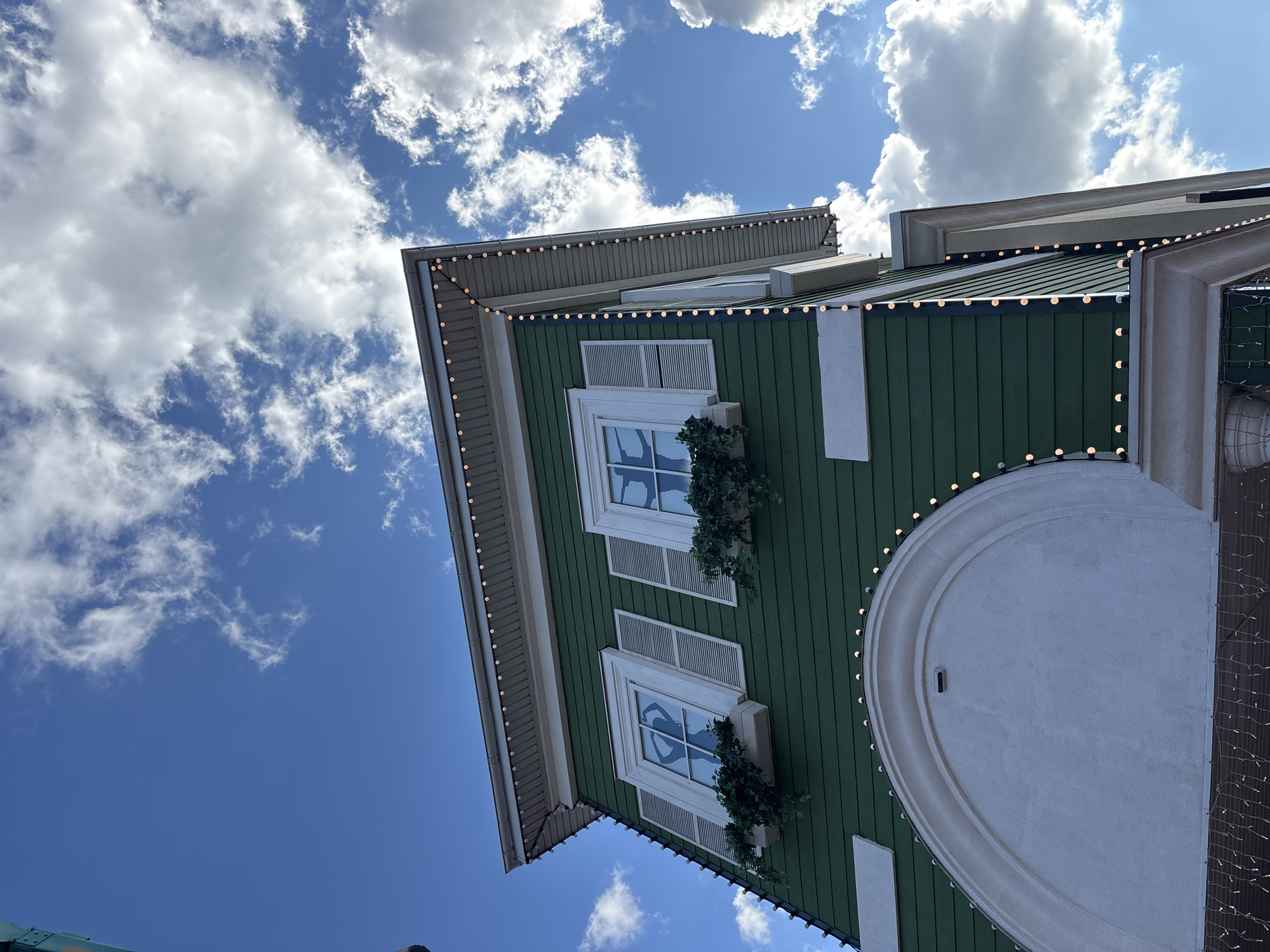 The image shows the upper facade of a green building, possibly a house or commercial establishment, against a bright blue sky with scattered white clouds. The building has white-trimmed windows with shutters and window boxes filled with greenery. A string of small, illuminated lights runs along the eaves of the roof and down the side of the building, suggesting a festive or decorative purpose. The architectural style appears somewhat whimsical, with an arched white element on the lower part of the visible facade. No people are visible in the image. The context of Pokrovskoe, Russia is not discernible from the visual information alone.