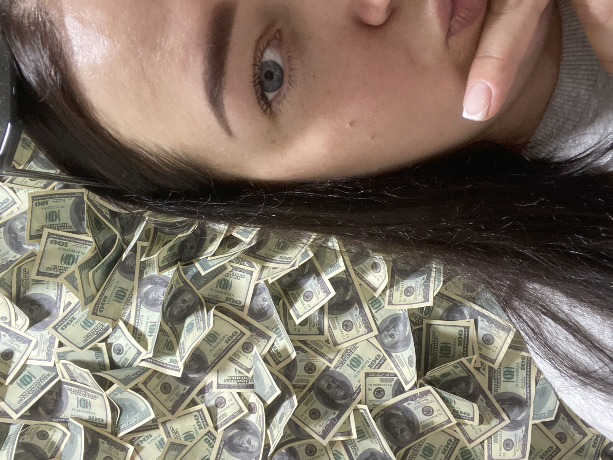 A woman with long, dark hair and light blue eyes is lying on a pile of US one hundred dollar bills. Her face is partially obscured by her hand near her mouth. The background consists entirely of the scattered currency. There is no evidence to suggest the location is in Sochi, Russia.