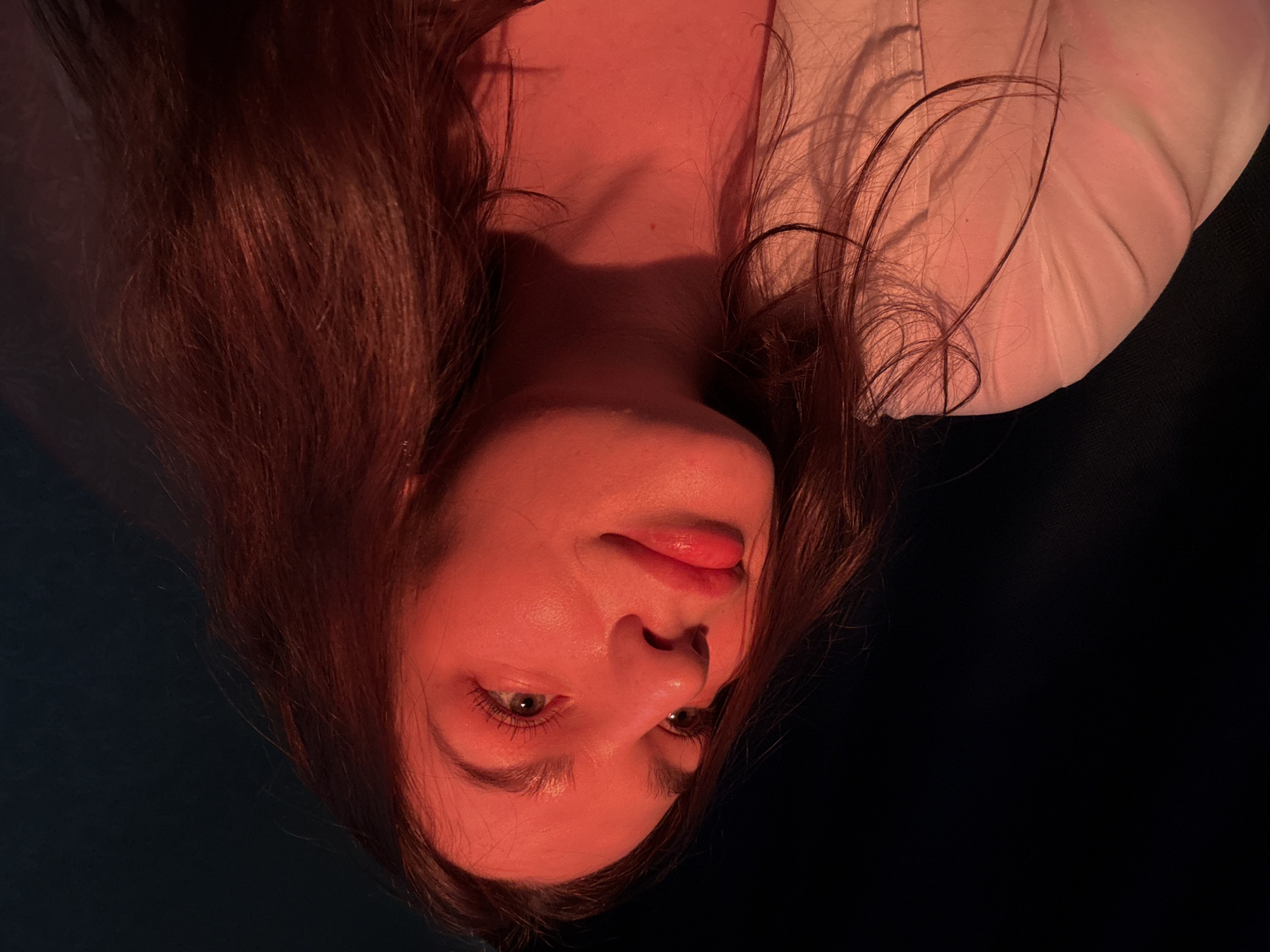 A close-up, upside-down view depicts a woman with fair skin, reddish-brown hair, and light-colored eyes. The subject is lit with a warm, reddish light, highlighting the contours of her face, neck, and hair. She appears to be wearing a light-colored garment. The background is dark and indistinct. The location is not discernible from the image alone.