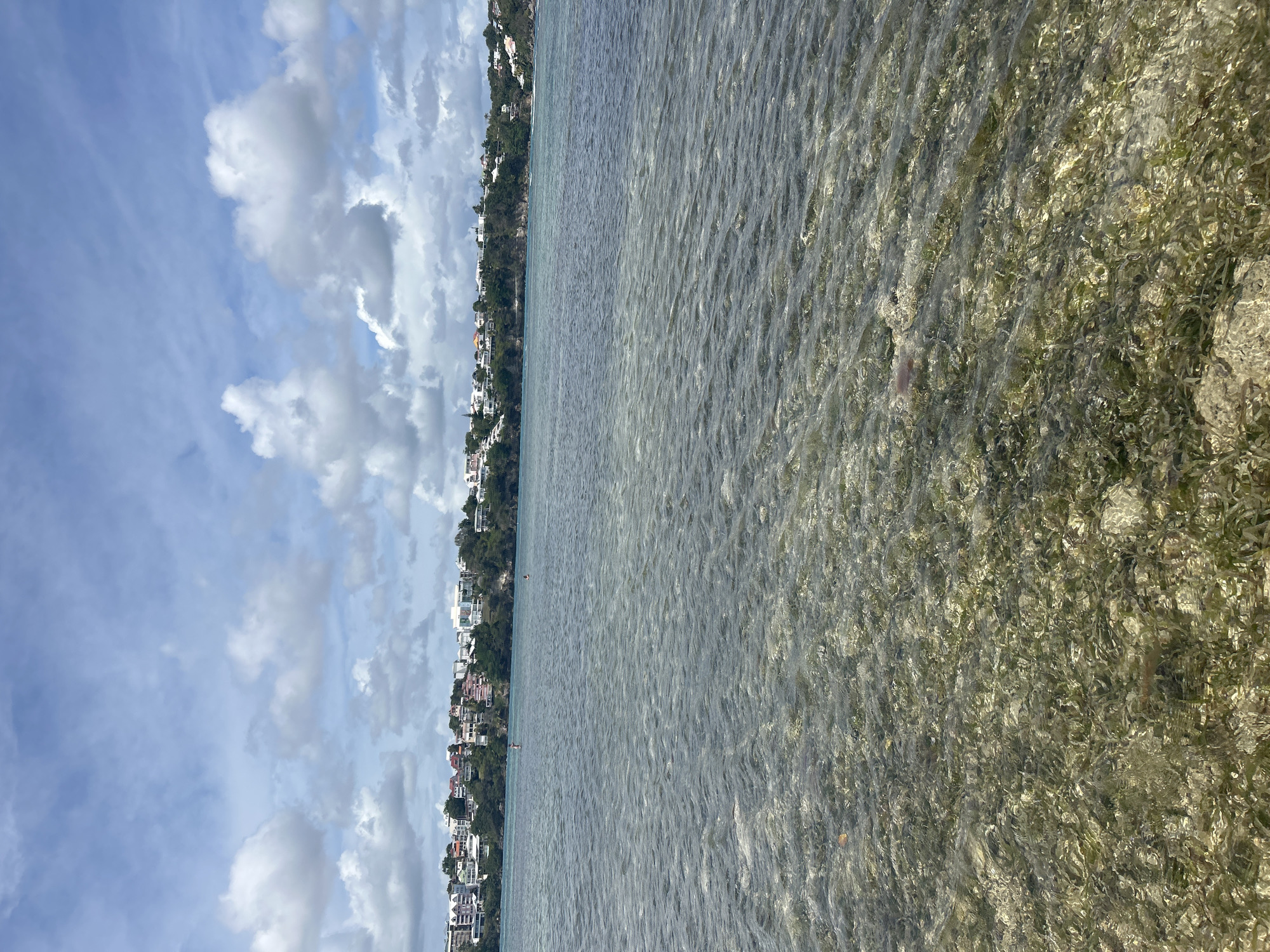 The image shows a coastal scene in Le Gosier, Guadeloupe, France. A body of water, possibly a shallow lagoon or bay, occupies most of the frame, with clear, slightly rippled water revealing a sandy bottom and some submerged vegetation. Along the shoreline, there are buildings and houses clustered together, backed by lush green trees and vegetation. The sky is partially cloudy with patches of blue visible through the clouds.