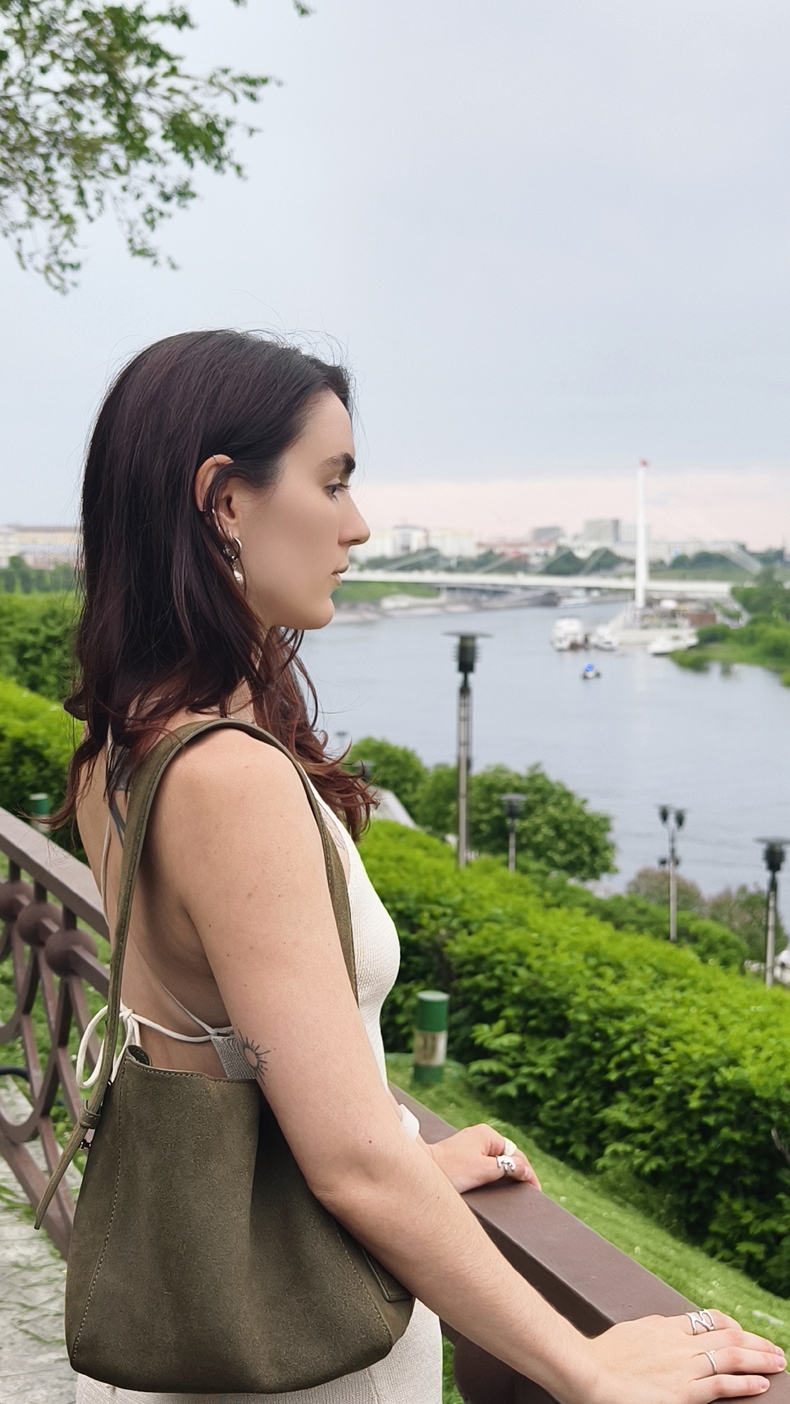 A young woman stands at a railing overlooking the Tura River in Tyumen, Russia. She has dark hair, fair skin, and is wearing a light-colored top. A green shoulder bag hangs from her left shoulder. Her hands rest on the railing, adorned with rings. In the background, the river flows with boats, and the cityscape is visible with buildings and a prominent white pylon. Lush green bushes line the embankment, adding to the park-like setting. The sky is overcast.