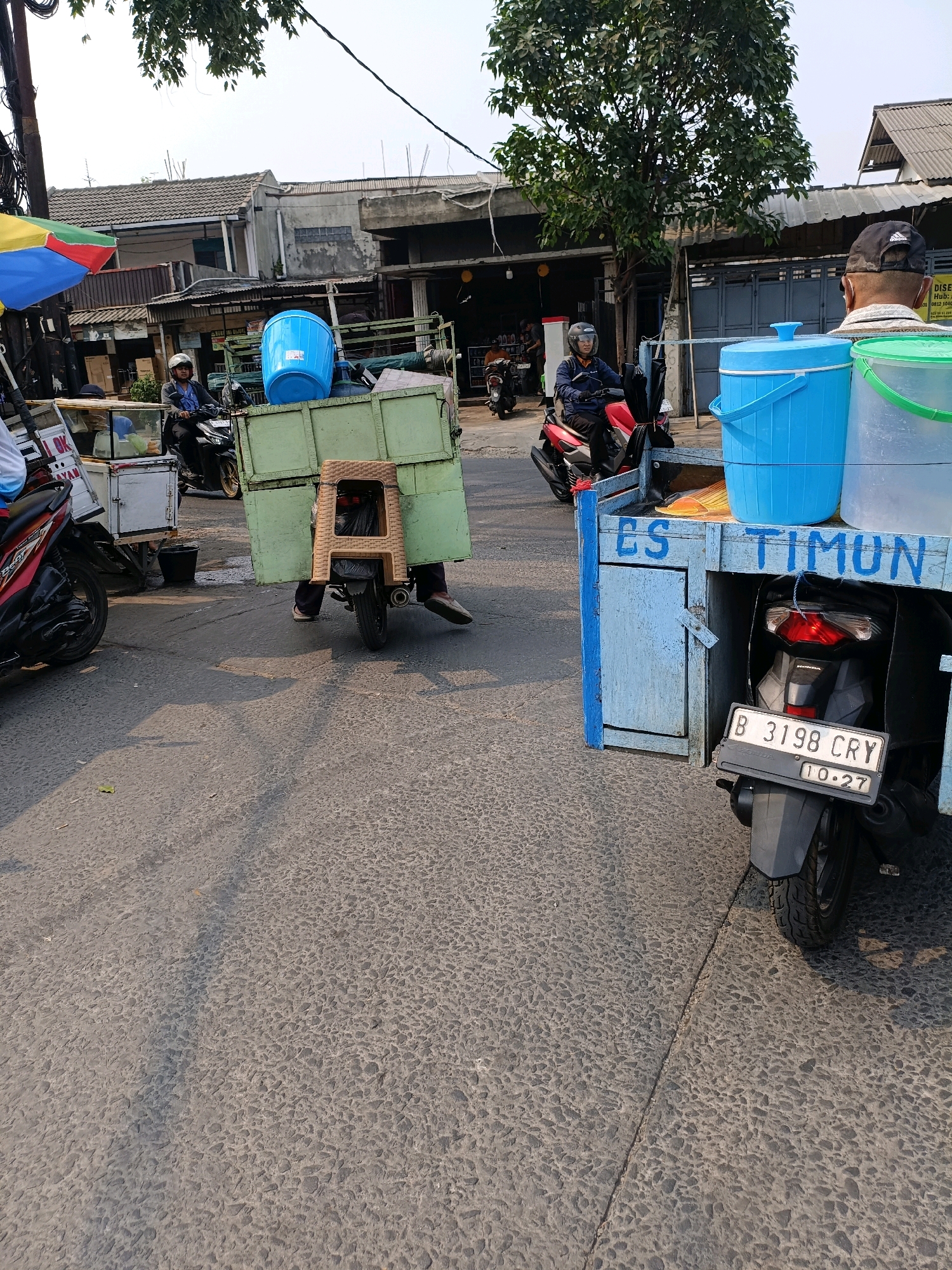 A street scene in Tangerang, Indonesia shows an ice vendor's cart being pulled by a motorcycle. The cart is a wooden structure painted blue with "ES TIMUN" painted on the side, indicating it sells cucumber ice. On the cart are two large blue tubs, likely for storing ice and drinks. A man on a red motorcycle is pulling the cart, wearing a blue jacket and helmet. To the left, another motorcycle and cart are visible. The street is paved and appears to be a commercial area with buildings in the background.