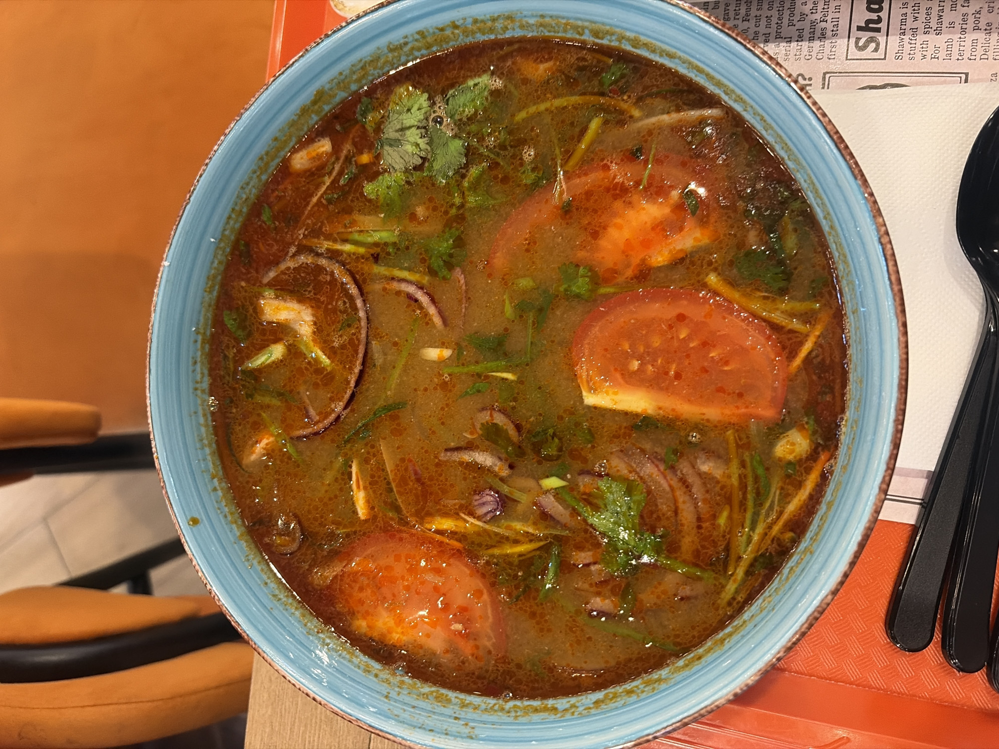 A top-down shot captures a steaming bowl of hearty soup, likely a spicy or sour variant, served on a light wooden table. The soup, presented in a vibrant blue ceramic bowl with a rustic, textured rim, features a rich, reddish-brown broth. Visible ingredients include several large slices of red tomato, translucent rings of red onion, and generous garnishes of fresh green herbs, possibly cilantro or parsley. Small bubbles on the surface suggest it was recently served and is still hot.
To the right of the bowl, a neatly folded white napkin holds black cutlery, likely spoons or forks, resting on an orange, textured placemat or tray. Partially visible in the upper right background is a piece of paper, possibly a menu or newspaper clipping, featuring text with the prominent headline "Sha" and the word "Shawarma." The surrounding text discusses shawarma, mentioning it is "stuffed with spices," and detailing its preparation with "lamb" and contrasting it with "pork." Other snippets of text are also visible, discussing a product "started by a Germany" and "Charles Feltn."
The background on the left reveals blurry orange upholstered seating and a light-colored floor, suggesting a casual restaurant or eatery setting. The overall scene depicts a prepared meal, possibly in Izhevsk, Russia, highlighting a robust-looking soup amidst a dining environment that also offers Middle Eastern cuisine.