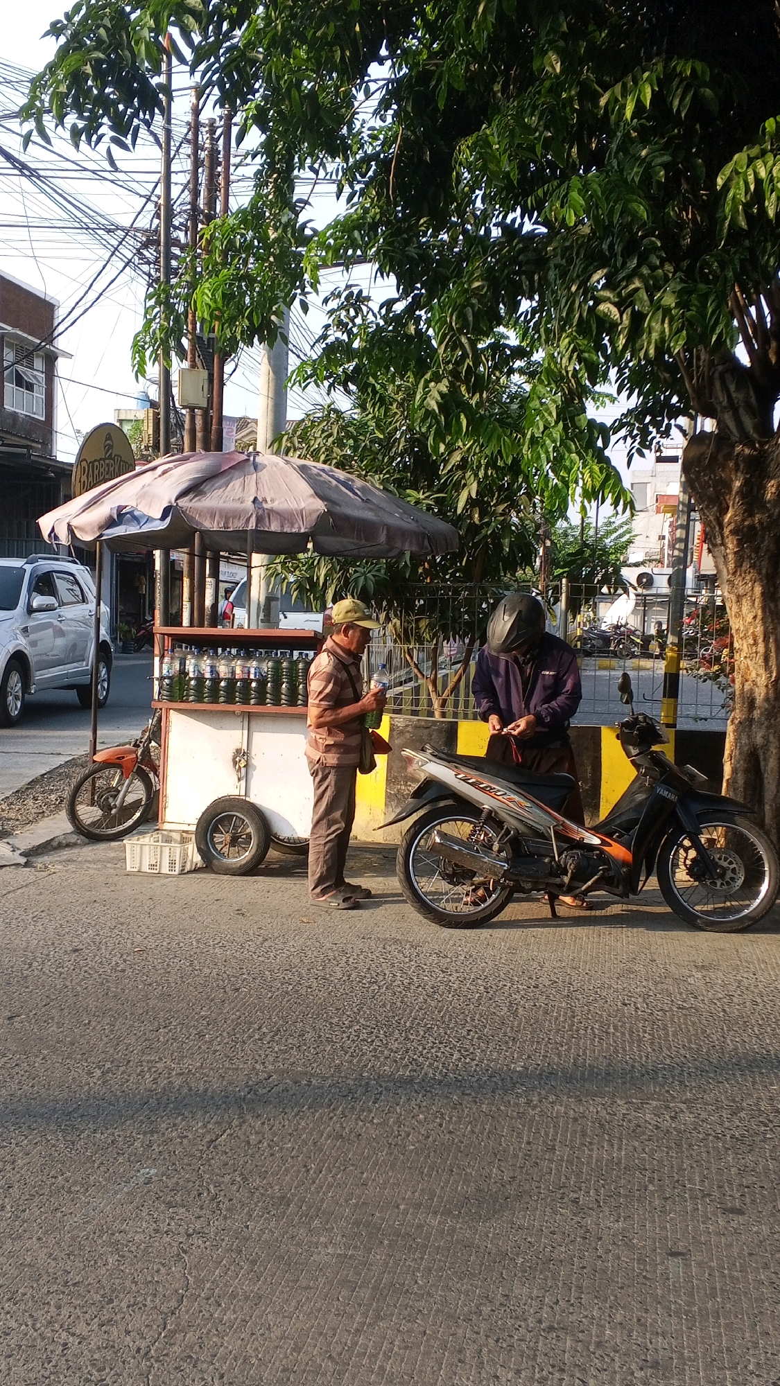 The image depicts a street scene in Semarang, Indonesia, with a focus on two individuals and a beverage cart. A vendor, an elderly man wearing a striped shirt, brown pants, a green cap, and sandals, stands beside a mobile cart filled with bottled beverages. He is holding a bottle of liquid. To his right, another man, wearing a dark jacket and a helmet, is interacting with the vendor, possibly making a purchase. A motorcycle is parked beside the second man. The cart is under a large umbrella and appears to be set up on the side of a road, with a white SUV visible in the background. The environment suggests an urban setting with trees and utility poles present. The lighting indicates it is daytime, likely morning or late afternoon due to the shadows.