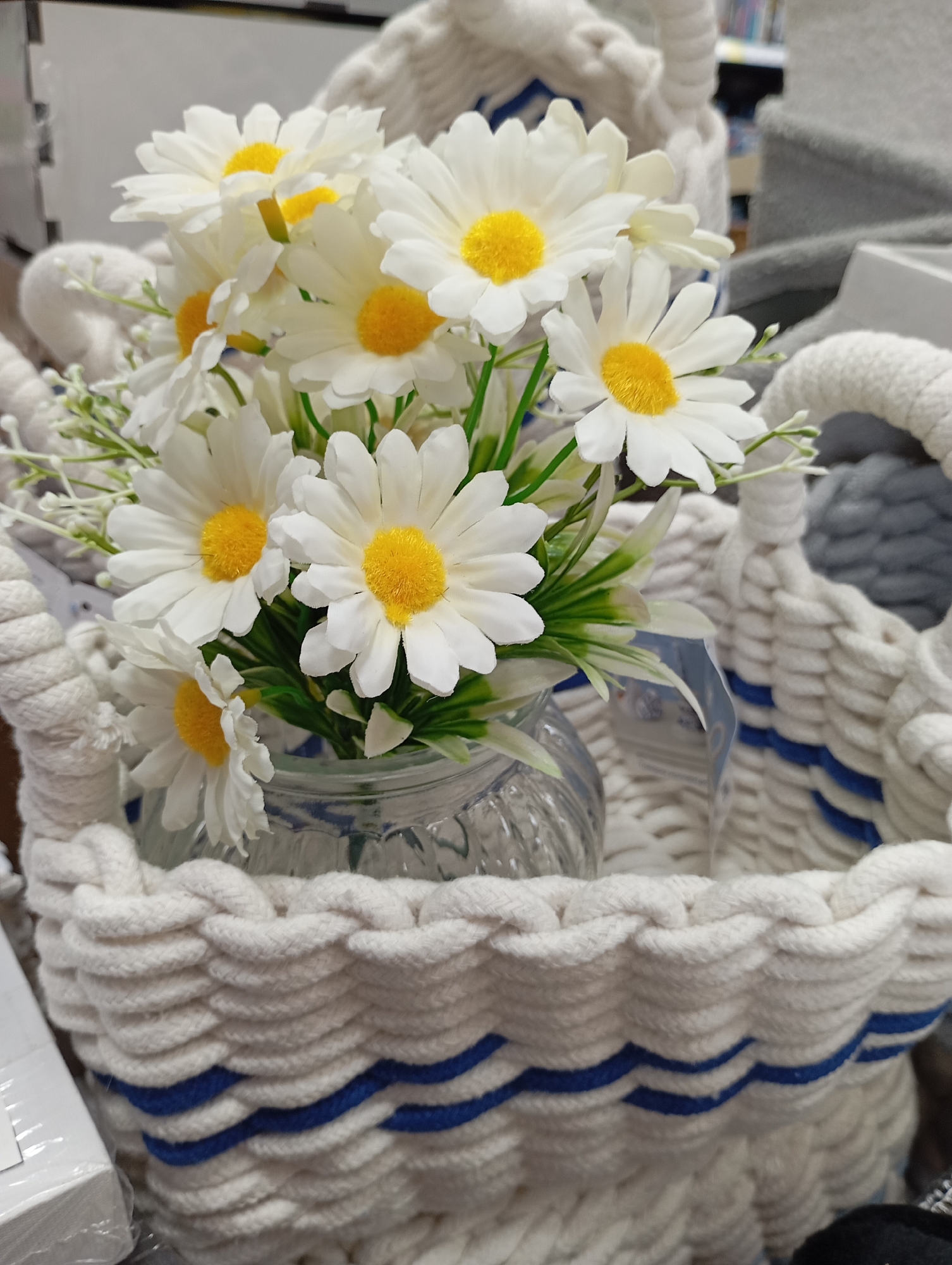 A high-angle, close-up shot shows a bouquet of artificial daisies arranged in a clear glass vase, nestled within a white rope basket adorned with blue stripes. The daisies are primarily white with prominent yellow centers, surrounded by green foliage and delicate white sprigs. The vase, partially obscured, is filled with water that is visible through the glass. The basket is intricately woven from thick white rope, creating a textured surface. Blue stripes are woven into the rope at regular intervals. In the blurred background, gray fabric and retail shelving are visible. The setting appears to be a retail store, possibly in the city of Unknown, Unknown.