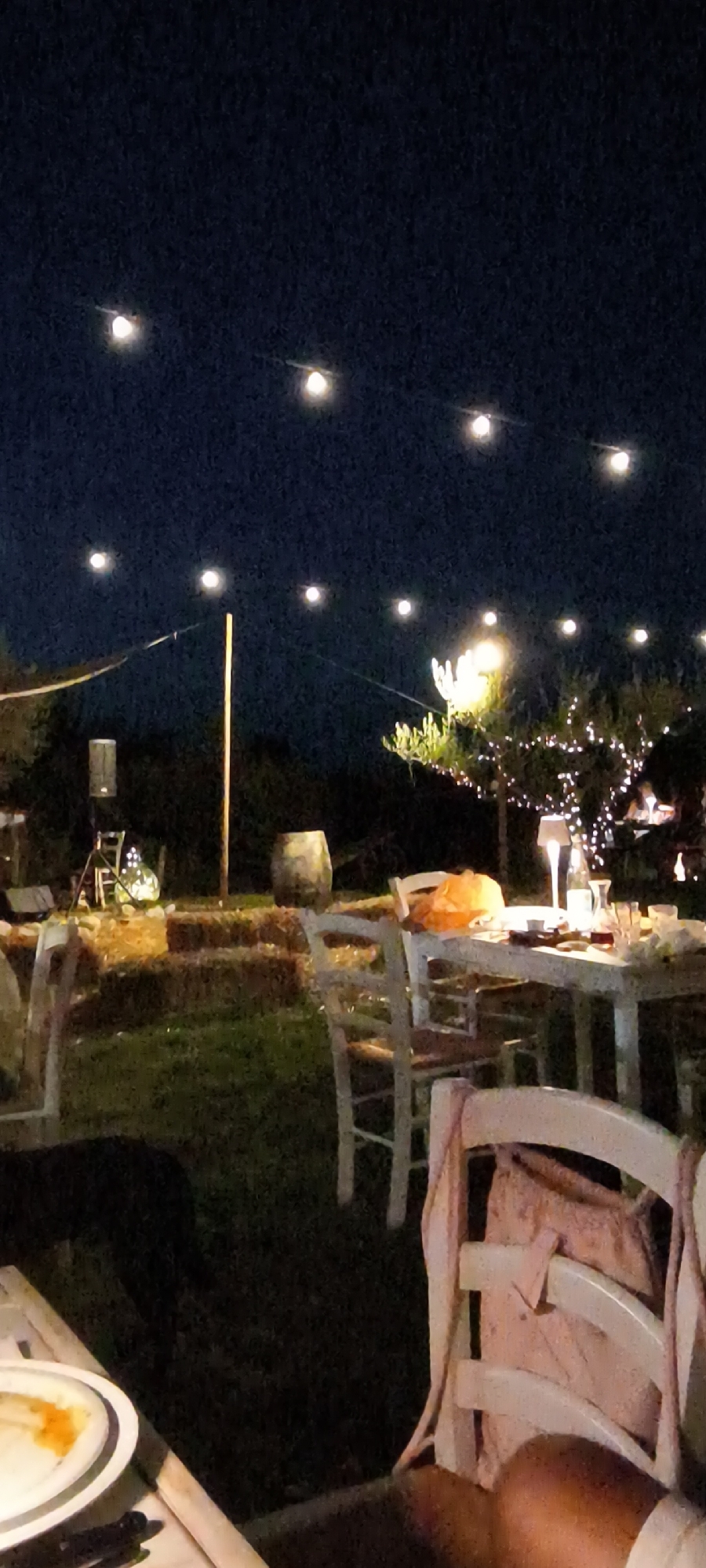 The image depicts an outdoor dining setup at night in the vicinity of Buriano, Italy. The scene is illuminated by strings of fairy lights suspended overhead and a tree adorned with lights in the background. In the foreground, several tables and chairs are arranged on a grassy area. One table in the mid-ground is set with tableware and a lamp. In the background, there are hay bales, a wooden barrel, and a speaker on a stand. The overall ambiance suggests a rustic, nighttime gathering or event.