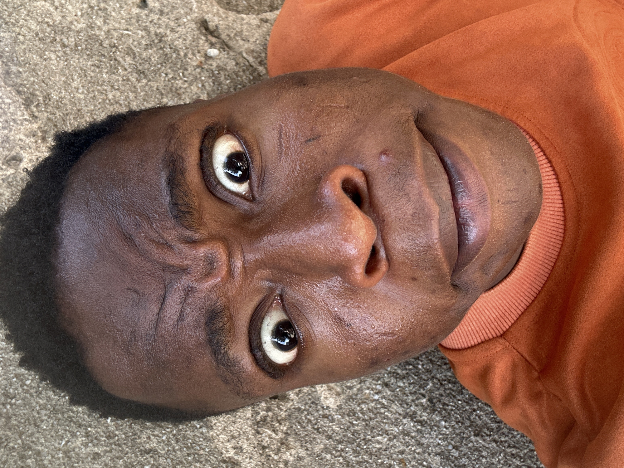 A person with dark brown skin, close-cropped dark hair, and an orange sweatshirt is lying on a rough, light gray surface, possibly concrete. The person's face is turned upward, with eyes wide open and eyebrows furrowed. There are some faint lines or marks on their forehead. The location is in the city of Unknown, Unknown.