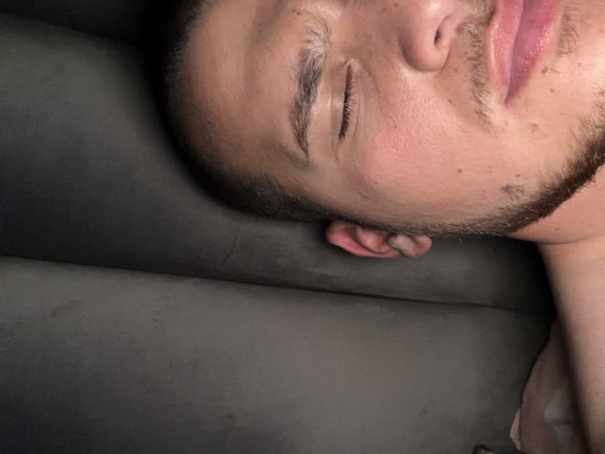 A close-up, angled shot shows a man sleeping on a grey upholstered surface, likely a sofa or bed. His face is prominent, displaying closed eyes, facial stubble, and fair skin. Part of his neck and shoulder are visible on the right. His ear rests on the cushion. The background is out of focus. The location is not discernible from the image.