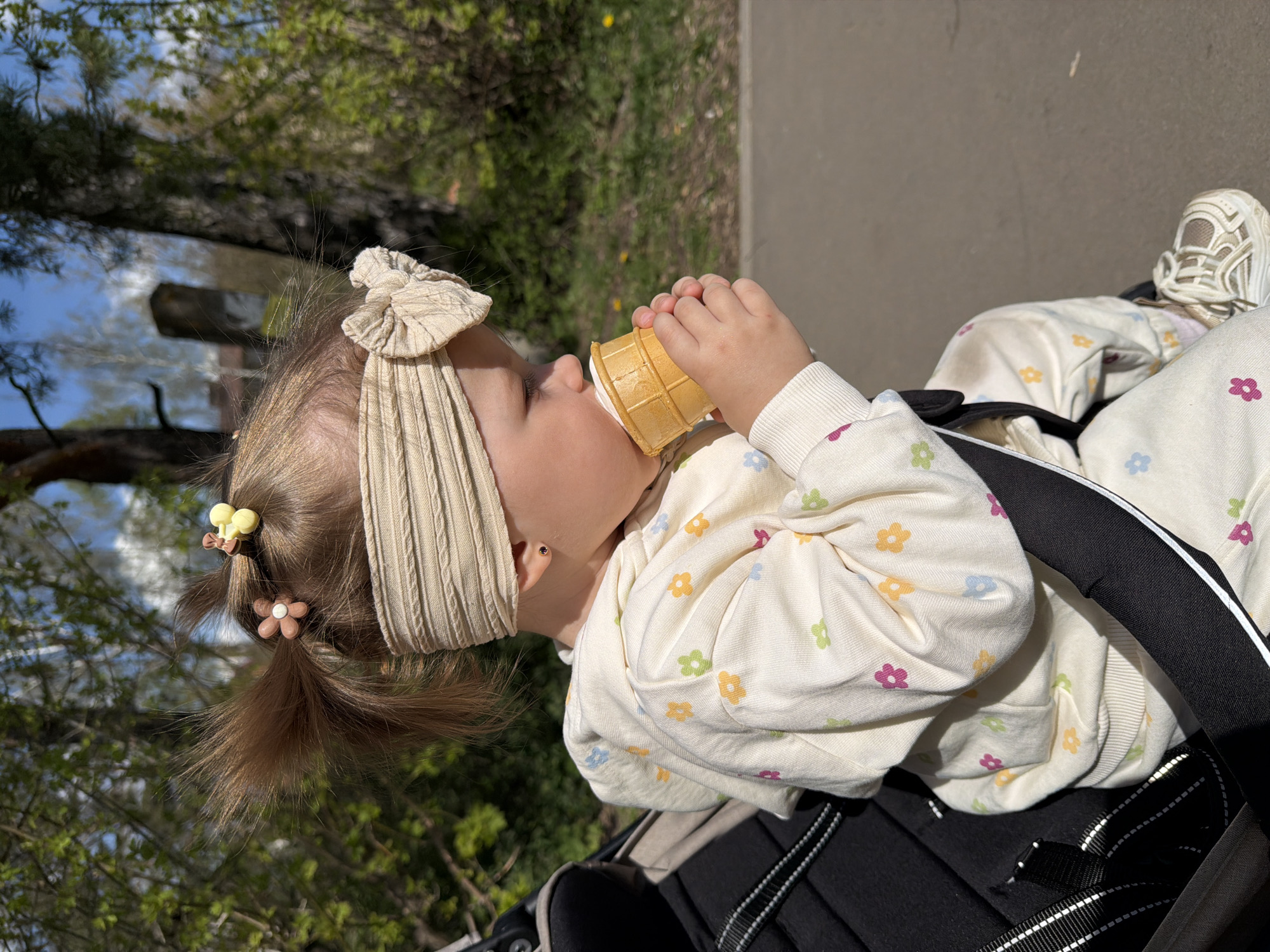 A young child sits in a stroller outdoors. The child wears a floral-patterned outfit, a beige ribbed headband, and hair ties with flower decorations. The child is holding and eating an ice cream cone. The stroller is black. The background shows trees and a pathway. The location is Reshetnikovo, Russia.