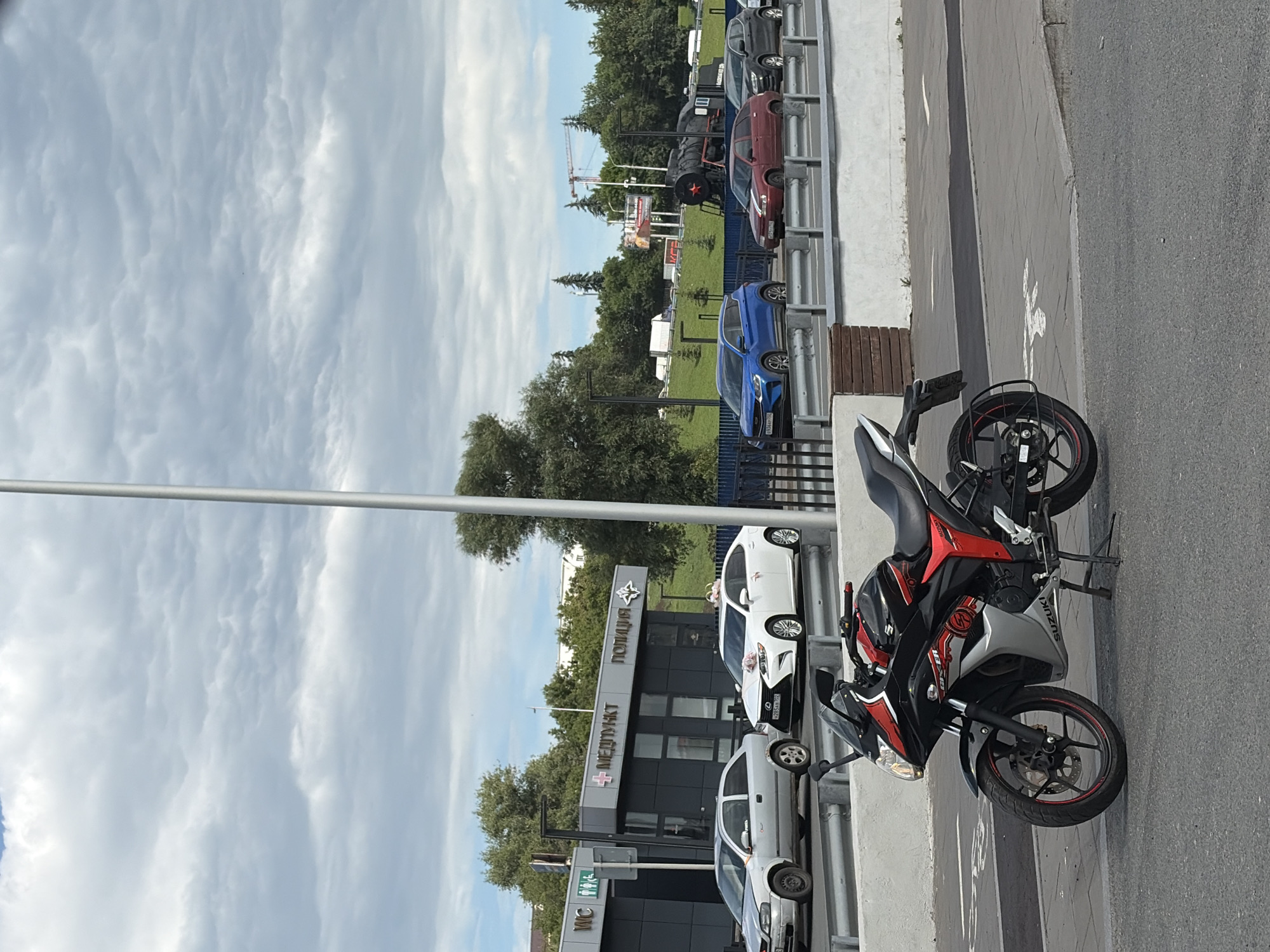 A black and red Suzuki motorcycle is parked on an asphalt road next to a concrete barrier. In the background, there is a line of parked cars, including a blue sedan, a red sedan, and a white car decorated with pink ribbons, suggesting a possible wedding. Behind the cars, there are trees and a building with signs that read "ПОЛИЦИЯ" (Police) and "МЕДПУНКТ" (Medical Point). The sky is cloudy, indicating overcast weather. The overall scene appears to be a street or parking lot in Ufa, Russia. The time of day is likely daytime, given the lighting.