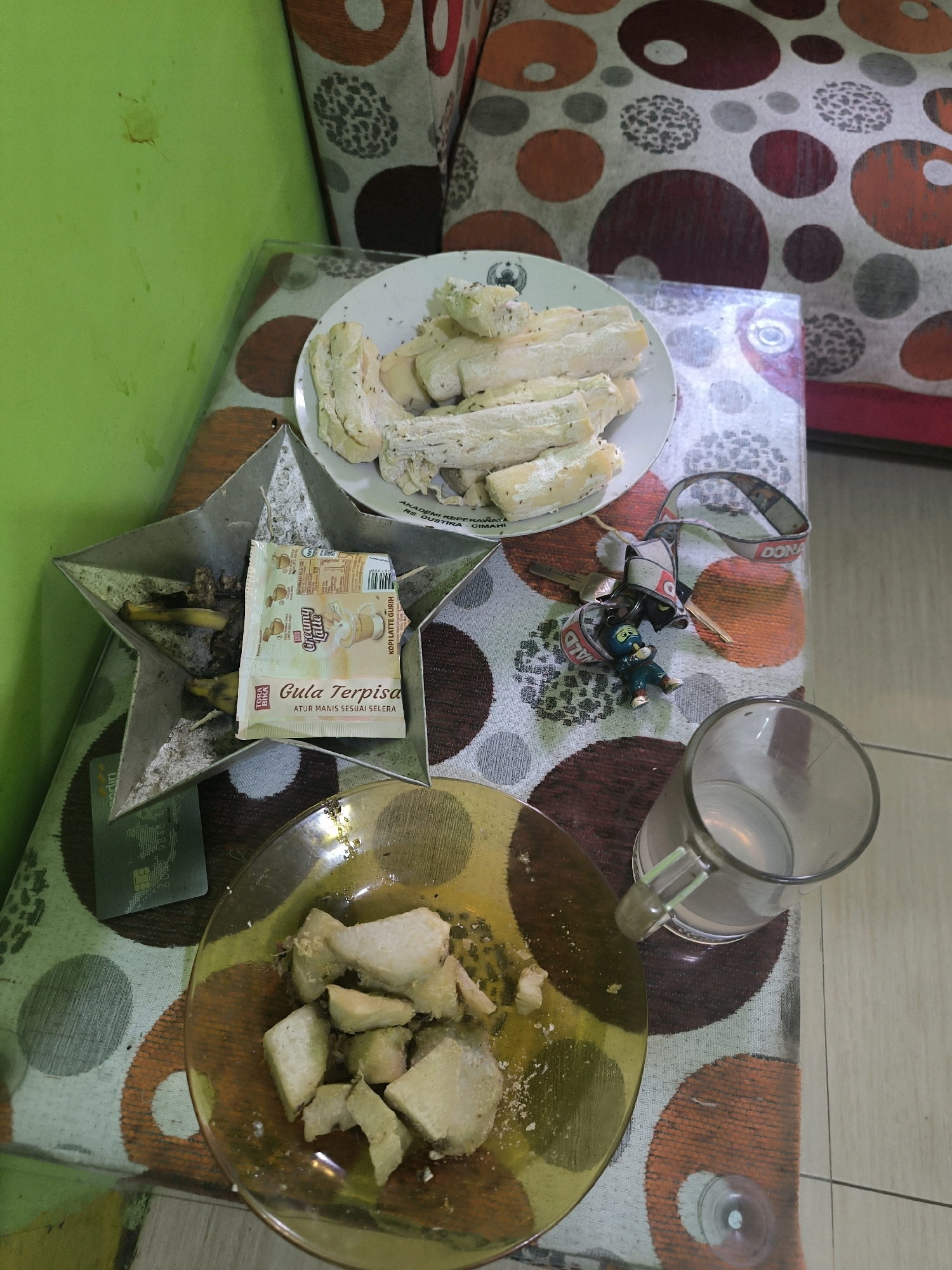 The image depicts a low-angle, eye-level view of a table in what appears to be a residential setting in Cilengkrang, Indonesia. The table is covered with a patterned tablecloth featuring circles of orange, brown, gray, and white.
On the table, there are several items of food and drink. To the left, a star-shaped ashtray contains what appear to be cigarette butts and two small packets of instant coffee. Above the ashtray, a plate holds several pieces of what seems to be steamed cassava, some of which are topped with a sprinkle of dark specks. To the right of the ashtray, a clear glass mug is partially filled with a light-colored liquid, possibly water. In the foreground, a yellow-tinted glass plate holds more pieces of steamed cassava, some of which appear to be seasoned with a yellow powder.
Among the food items, a lanyard with a small keychain figure is visible, along with a key. A credit card is also present, positioned on the green surface to the left of the table. The background shows a green wall and a section of furniture with a similar circular pattern to the tablecloth. The overall impression is that of a casual, domestic scene, possibly a meal or snack break.