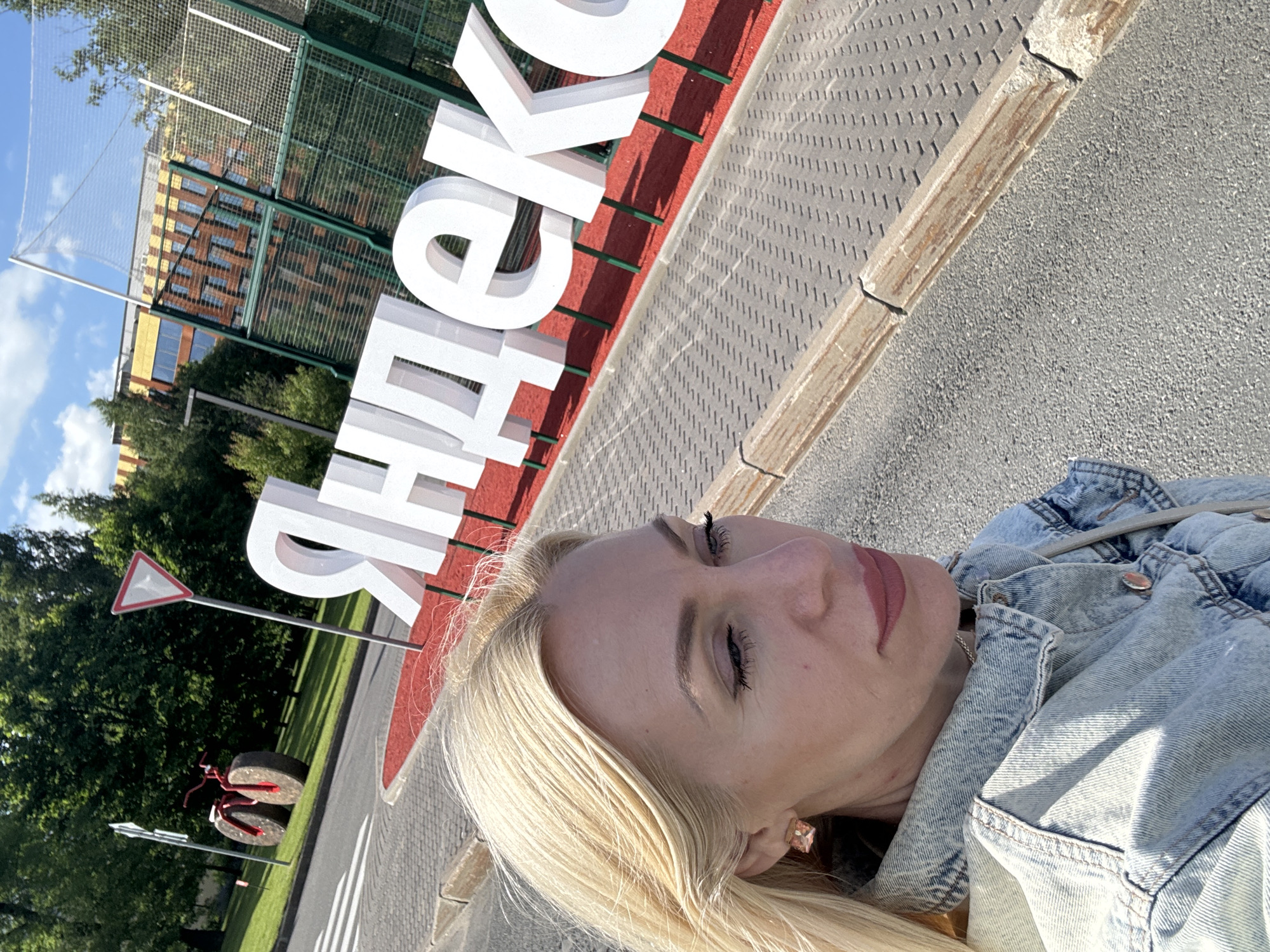 A blonde woman, wearing a light blue denim jacket, is captured in a close-up selfie. Her eyes are looking down and to the right, with subtle makeup emphasizing her lashes and lips. In the background, large white letters spelling "Яндекс" (Yandex) are visible against a backdrop of trees and a modern building with yellow and brown accents. A green sports net enclosure is also present. To the left, a unique red bicycle sculpture with oversized wheels is situated on a grassy area next to a pedestrian crossing and a yield sign. The scene appears to be an outdoor corporate campus or public space. The Yandex logo suggests the location is likely the Yandex headquarters or a branch office.