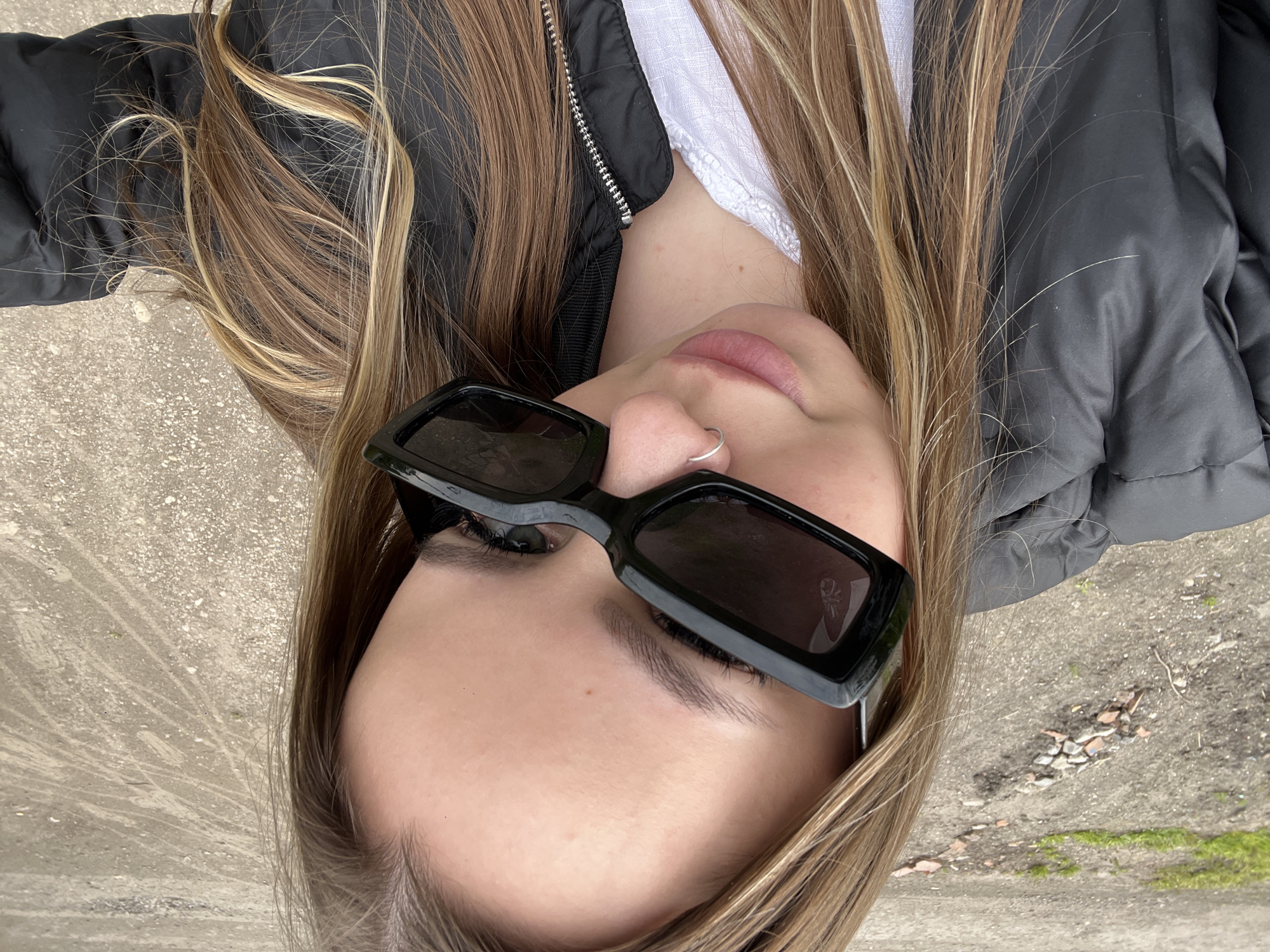 A young woman with long blonde and brown hair is lying on what appears to be a concrete surface outdoors. She is wearing black rectangular sunglasses, a white shirt, and a black puffer jacket. A silver nose ring is visible. The background includes a rough, uneven concrete surface with some debris and possibly grass or moss. The photograph is taken from an overhead perspective. Based on the information provided, the location is Port-Katon, Russia.