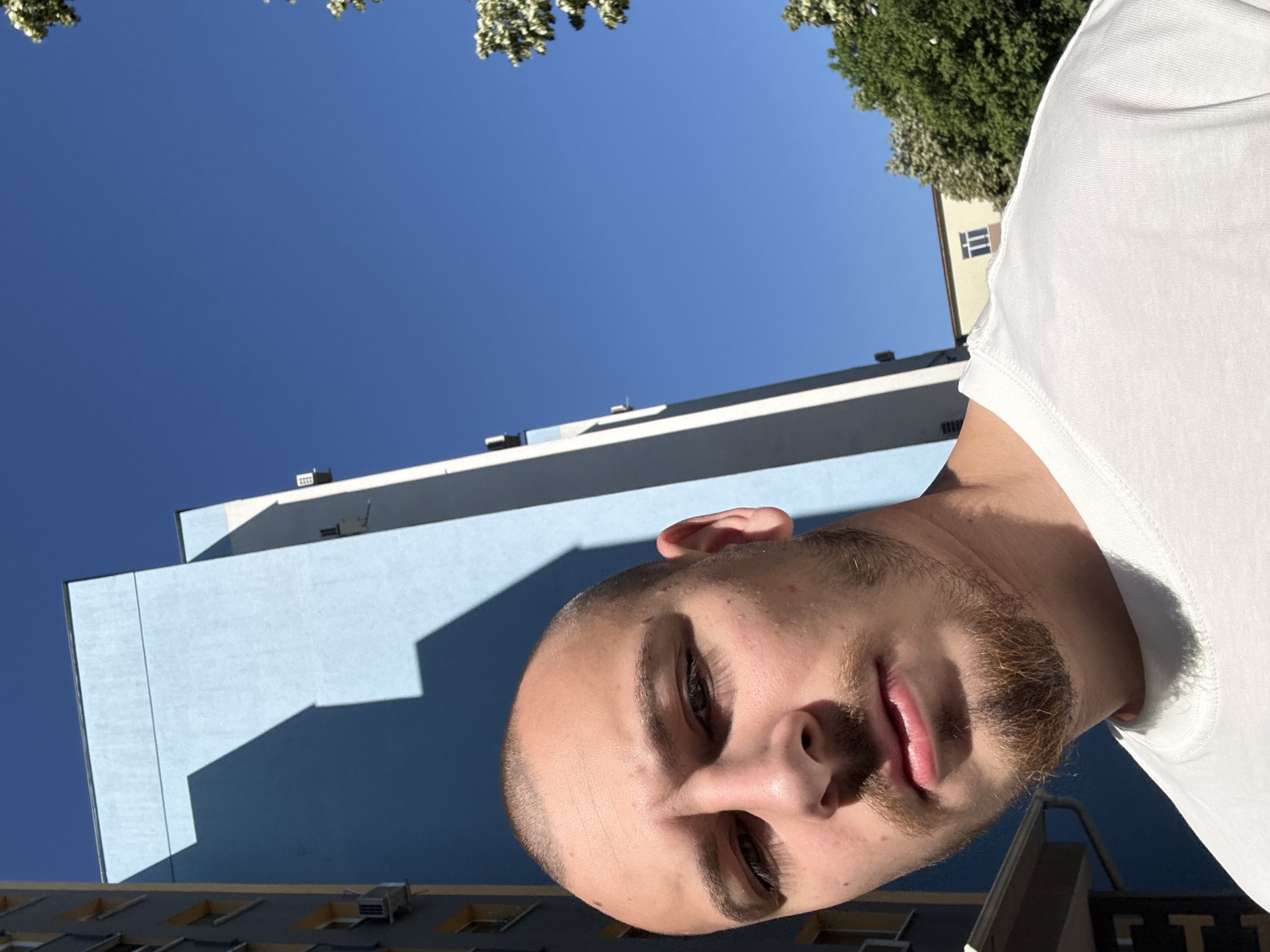 The image depicts a close-up of a young man with a shaved head and a light beard, wearing a white t-shirt. He is positioned in the foreground and appears to be taking a selfie. Behind him, a blue building with a distinct geometric shadow cast by the sun dominates the background. To the right, lush green trees with white blossoms and a clear blue sky are visible. A portion of a gray building with yellow window frames is present on the far left. The scene suggests an outdoor urban environment, likely in Stara Zagora, Bulgaria, given the architectural style and vegetation. The lighting indicates it is daytime with strong sunlight.