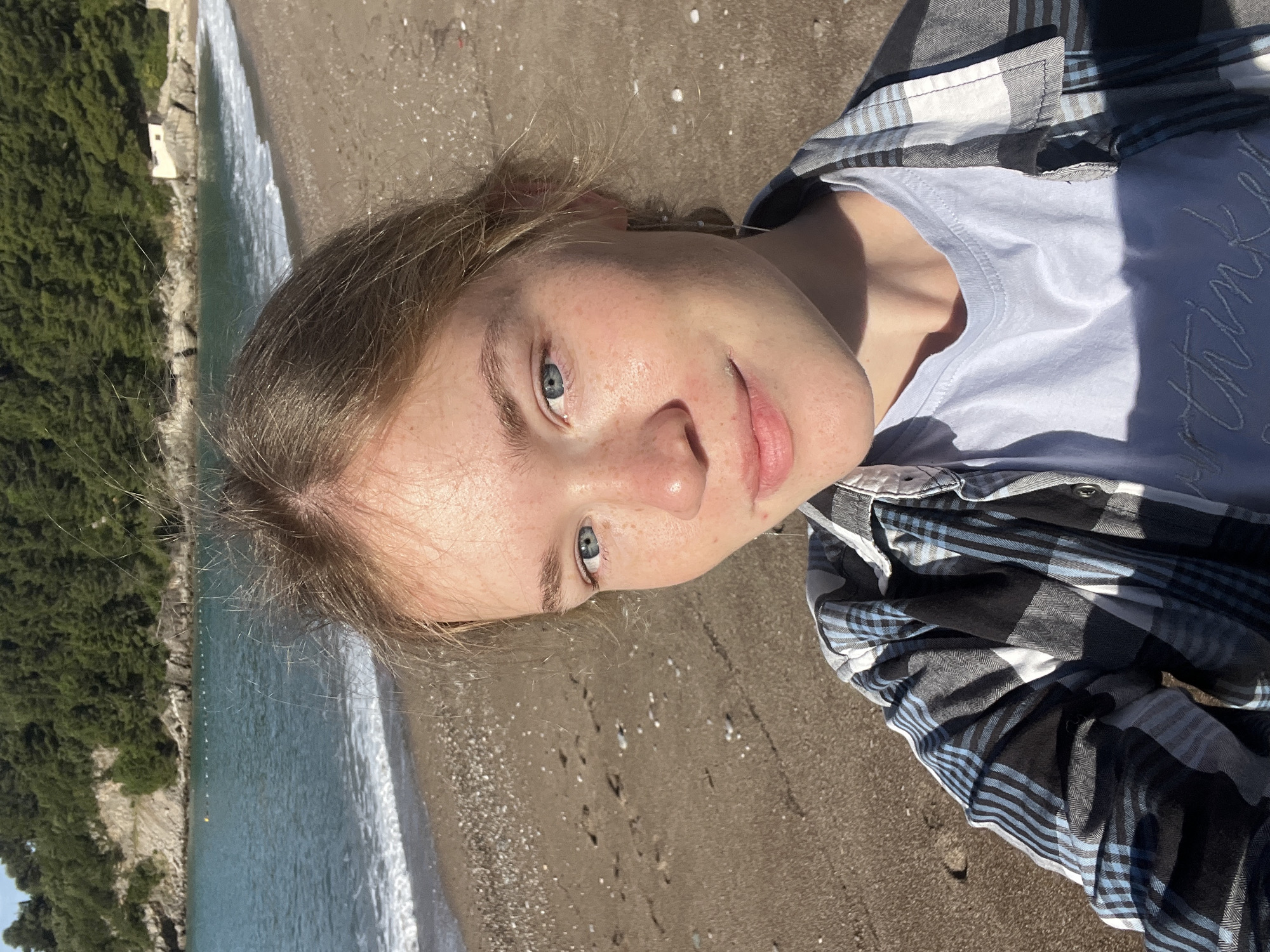 A fair-skinned young adult with blue eyes and freckles is photographed from a high angle on a beach in Bečići, Montenegro. The individual is wearing a light blue t-shirt covered by a black and blue plaid flannel shirt. They are positioned on the sandy beach, which is partially wet from the receding waves. Behind the beach, there is a rocky shoreline covered in dense green foliage and buildings. The sea is blue-green. The photograph has natural sunlight creating shadows and highlights.