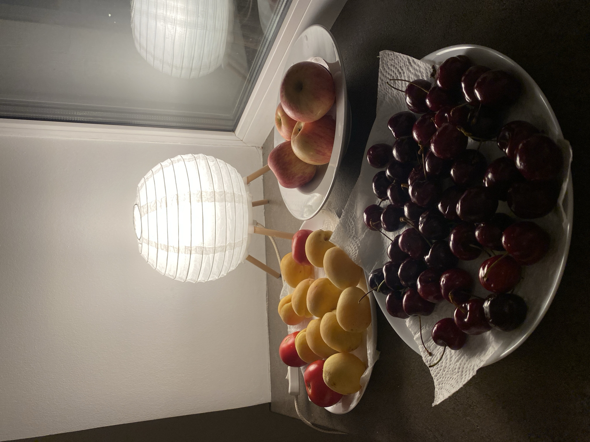 A still life photograph features three white plates filled with fruit, placed on a dark countertop. A white paper lantern lamp with a wooden base illuminates the scene from the left, casting a warm glow. To the right, a window reveals a dimly lit outdoor scene, with a reflection of another paper lantern visible in the glass.
The first plate, positioned on the left, holds several red tomatoes and yellow apricots. The second plate, centrally located, is filled with more yellow apricots. The third plate, in the foreground, is piled high with dark red cherries.
The image does not contain any information to suggest a location in Moscow, Russia.