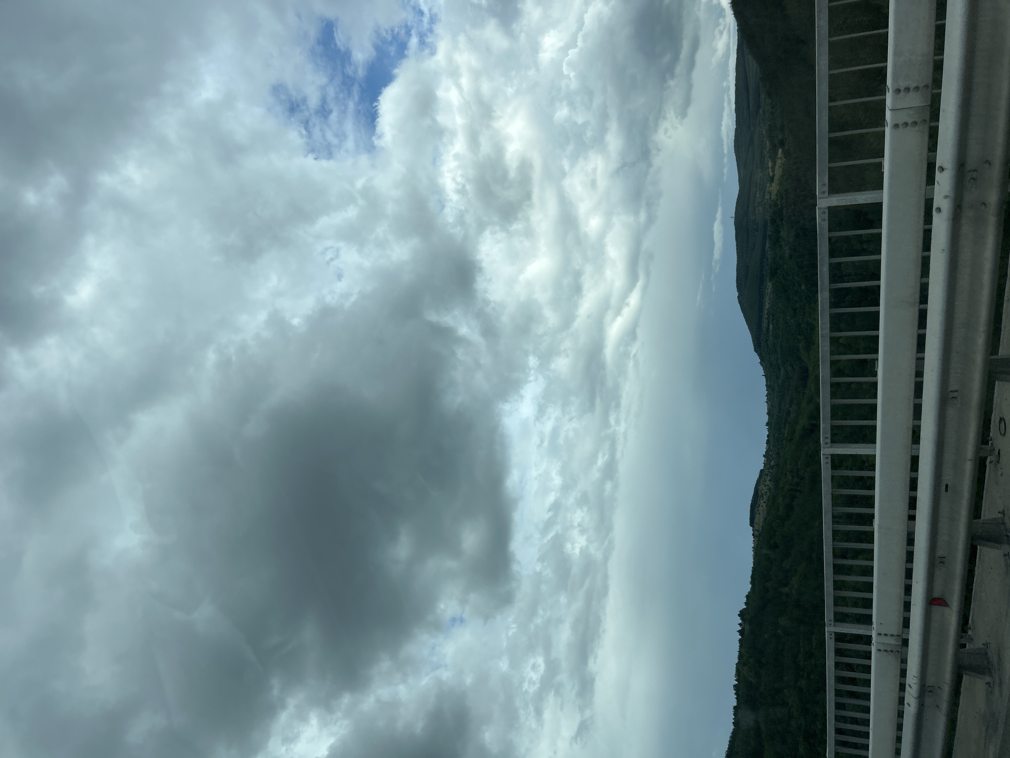 The image, taken with a vertical orientation and rotated 90 degrees clockwise, captures a broad sky with a forested mountainous landscape and a road barrier.
The sky occupies the majority of the frame, dominated by a dynamic interplay of clouds. Large, voluminous white and grey clouds are prominent, suggesting an active weather system. Patches of clear blue sky are visible, particularly towards the top left of the original image (which would be the upper left of a correctly oriented landscape image). Some clouds appear denser and darker, while others are brighter and more diffused, creating variations in light and shadow.
Below the cloudscape, a range of green, forested mountains or hills stretches across the middle distance. The vegetation appears dense, indicating a natural, perhaps rugged, environment. The sky above the distant hills transitions to a lighter, somewhat hazy blue.
In the foreground, along the right side of the image, a metallic guardrail or bridge railing is clearly visible. It consists of multiple horizontal bars and vertical supports, typical of roadside or bridge infrastructure. The metal appears to be silver or light grey, with visible bolts and structural elements. A small red reflector is barely visible on one of the lower bars of the railing.
The scene depicts a daytime setting with partly cloudy to mostly cloudy weather. There is no explicit activity taking place, but the presence of the railing suggests the photo was taken from a vehicle on a road or bridge. The location context is Dolno Varshilo, Bulgaria. There is no visible text in the image.