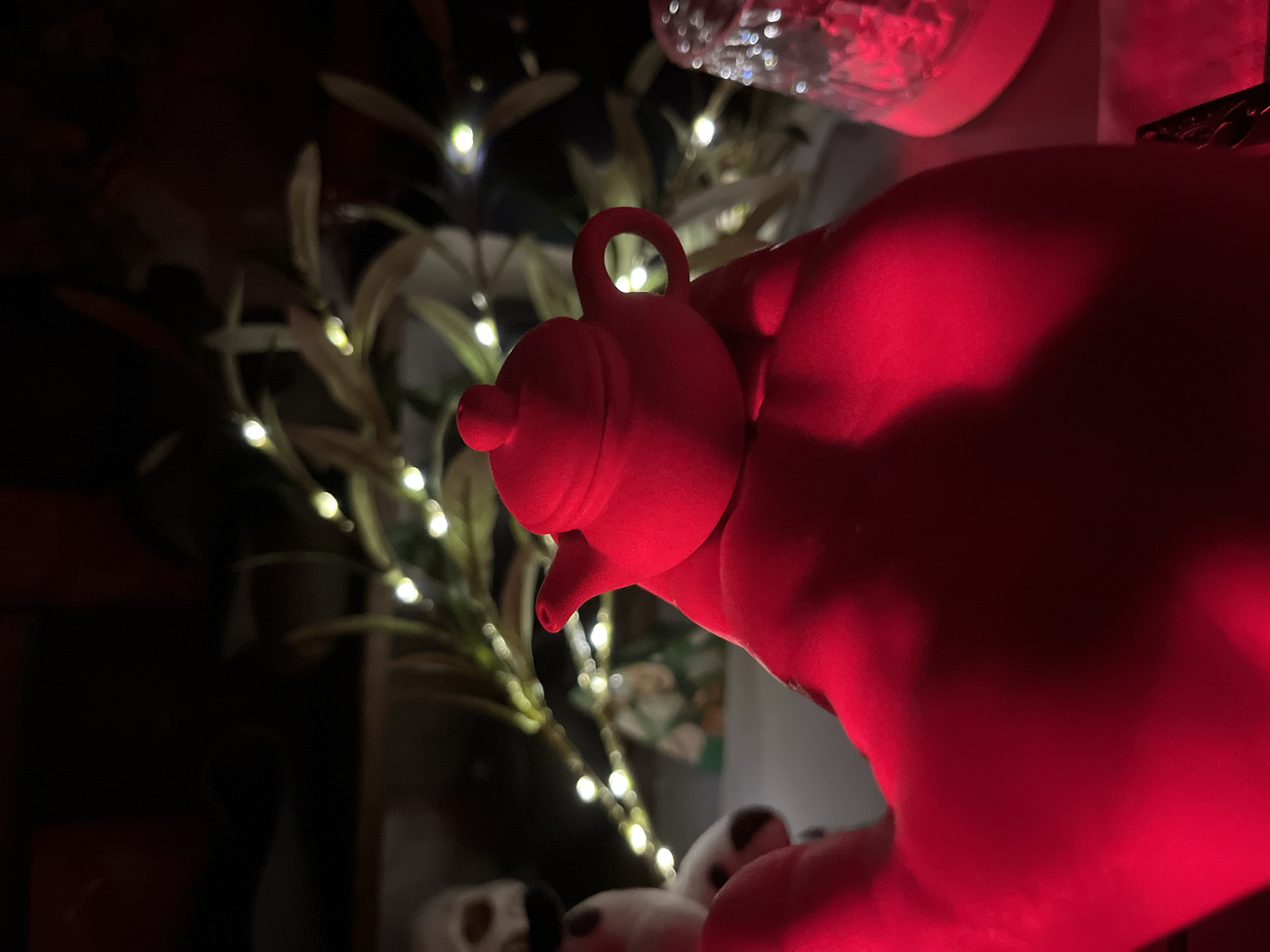An extreme close-up, red-lit shot shows a person's hand holding a miniature red teapot. The teapot has a rounded body, a lid with a small knob, and a handle. The background is dark with blurred details of plants and a string of small, bright lights. To the left of the teapot, partially visible, are what appear to be two white objects with dark spots, possibly decorative figures. To the right, a blurred object with a patterned surface is illuminated by the red light. The lighting creates deep shadows and highlights on the hand and teapot, emphasizing their form. The scene appears to be indoors, possibly a dimly lit room decorated with string lights.