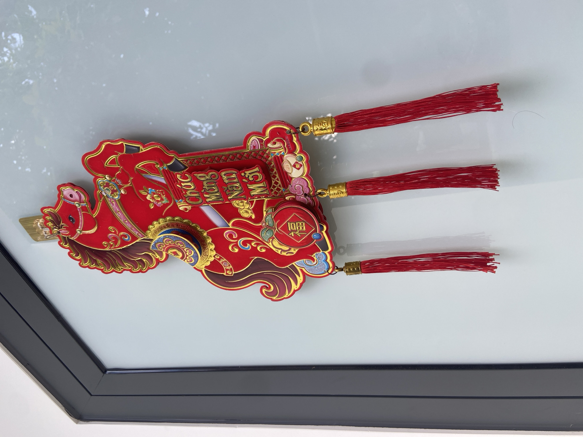 A red and gold Chinese New Year decoration in the shape of a horse with tassels hangs on a frosted glass door in Phu Thuy Ward, Vietnam. The horse is detailed with elaborate designs and the text "Chúc Mừng Năm Mới" (Happy New Year) is visible on a plaque attached to its saddle. A small red ornament with the Chinese character "福" (fortune) is also present. The door is framed by a dark grey border. Sunlight casts shadows on the surface of the door, suggesting it is daytime.