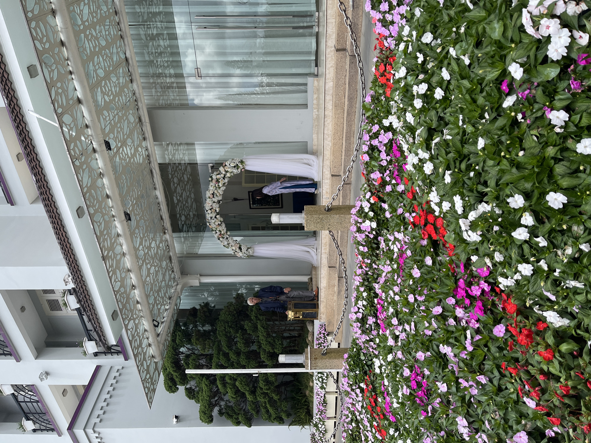 An elderly Vietnamese man stands near the entrance of a modern building, likely a hotel or resort, in Da Lat, Vietnam. The entrance features a contemporary design with glass doors and an elaborate, leaf-patterned awning. A floral archway adorned with white and pink flowers is positioned over the entryway, with sheer white fabric draped on either side. In the foreground, a vibrant bed of flowers in shades of pink, red, and white creates a striking visual contrast with the building's architecture. The man, wearing a dark jacket and trousers, stands beside a gilded chair, his posture suggesting he might be waiting or observing. Further inside the entrance, a young woman in traditional Vietnamese attire is partially visible, facing away from the camera. The overall scene captures a moment of serene elegance, blending natural beauty with modern design in the cool climate of Da Lat.