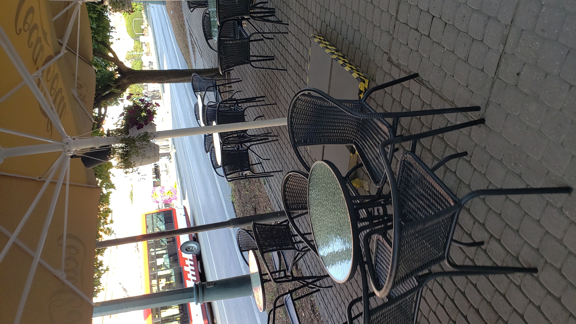 The image shows an outdoor seating area of a cafe with tables and chairs, situated on a cobblestone pavement. In the background, a red and white bus is visible driving on a street, indicating an urban environment. The seating area is partially covered by a large yellow umbrella. There are no people visible in the image. The scene appears to be a typical street-side cafe setting. The image is taken from a slightly elevated angle, looking down at the tables and chairs. The lighting suggests it is daytime, possibly late afternoon or early morning due to the shadows. The cafe appears to be empty. The specific location in Prešov, Slovakia, is not definitively identifiable from the image alone, but the presence of a bus on a city street is consistent with an urban setting.