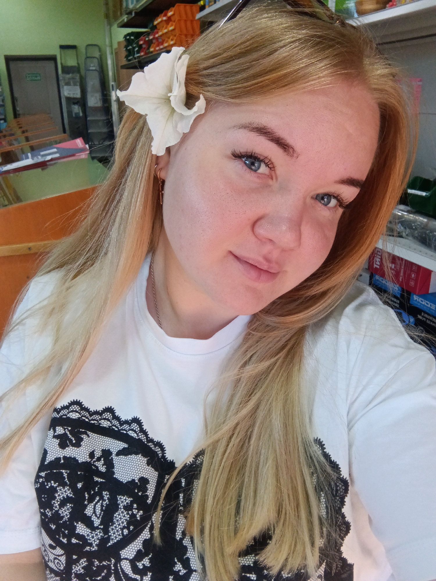 A close-up, selfie-style photo shows a young woman with blonde hair and blue eyes. She is wearing a white t-shirt with a black lace design on the front and a white flower tucked into her hair above her left ear. She is also wearing a gold earring in her left ear and a thin gold chain around her neck. The background appears to be an indoor setting, possibly a store or workshop, with shelves of products, ladders, and a green wall visible. The lighting is bright and natural. The image was taken in the city of Fokino, Russia.