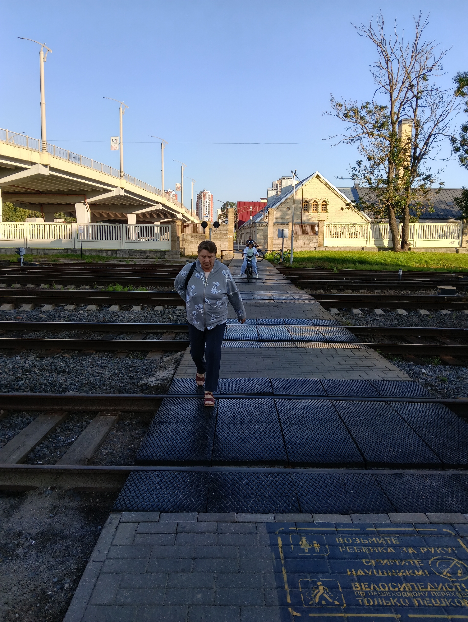 The image captures a pedestrian crossing over railway tracks in a city environment. In the foreground, a woman with short brown hair, wearing a grey patterned jacket and dark pants, is walking across the tracks on a textured surface. She is wearing sandals. Behind her, on a separate set of tracks, another person is riding a scooter. The background shows a low, light-colored building with arched windows, a white fence, and trees. An elevated road or overpass is visible in the upper left of the image, with streetlights. The sky is clear and blue. A sign with yellow lettering on the pedestrian walkway in the foreground displays pedestrian safety instructions, including icons for holding a child's hand, and a symbol for cyclists. The overall scene suggests a functional urban area with railway infrastructure and pedestrian access. The mention of Hrodna, Belarus is not visually ascertainable from the provided image.