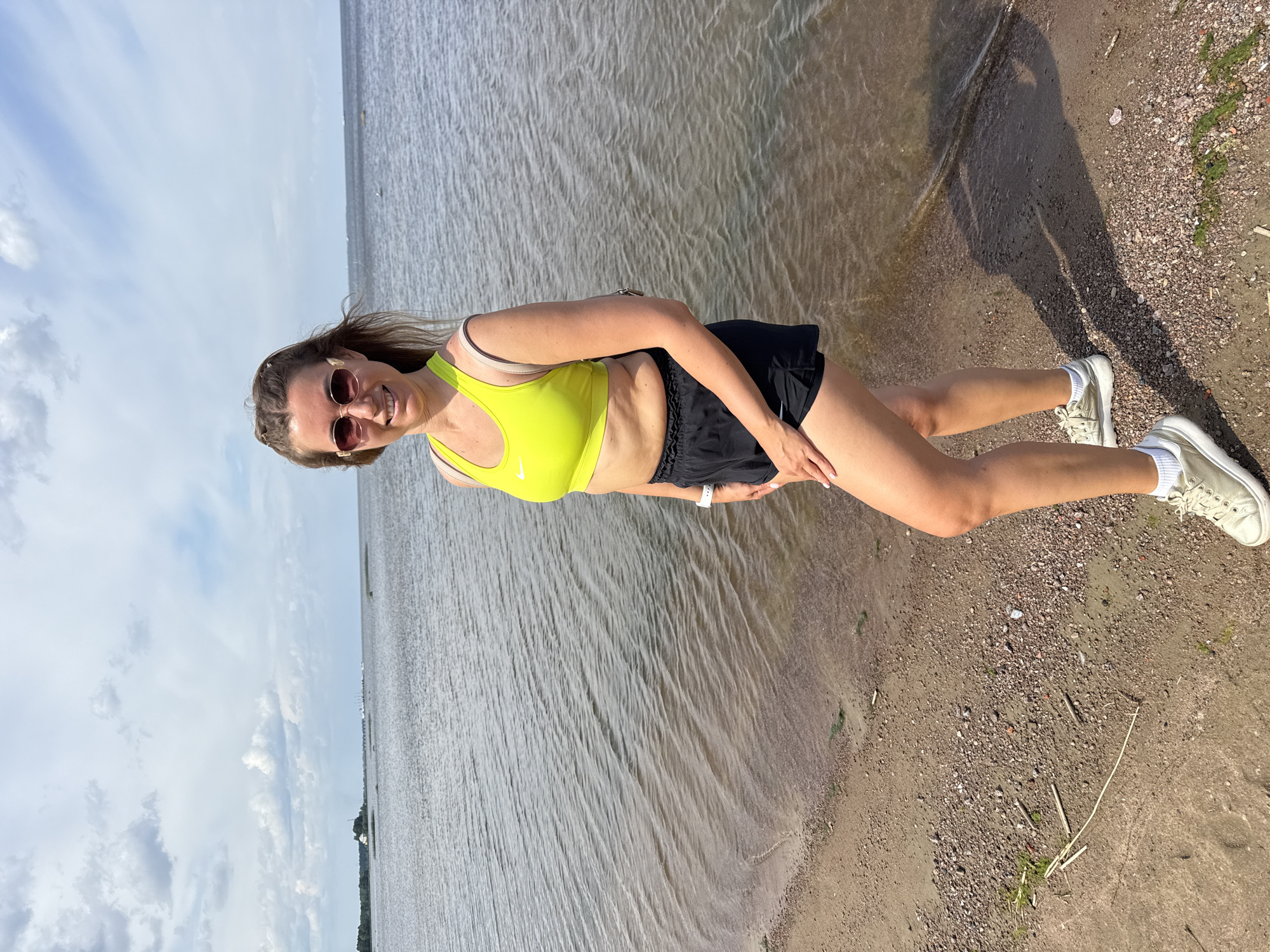 A female individual stands on a pebbled and sandy shore facing the camera, next to a body of calm, dark gray water under a partly cloudy sky. She has medium-long, light brown hair, wears dark, gold-toned framed sunglasses, and smiles slightly. She is dressed in a neon yellow Nike sports bra, black athletic shorts, and light-colored sneakers with white socks. A white smartwatch is visible on her left wrist, and a light-colored bag strap crosses her right shoulder. Her left hand rests on her left thigh. The shoreline in the foreground shows various small stones, sand, and patches of dark green algae. In the midground, the water extends to a distant horizon. To the far left, a distant landmass features trees and a small white building. Further along the horizon, several industrial or port structures and a faint bridge are visible. A shadow of the individual is cast on the sand to her right. The location is in the city of Unknown, Unknown.