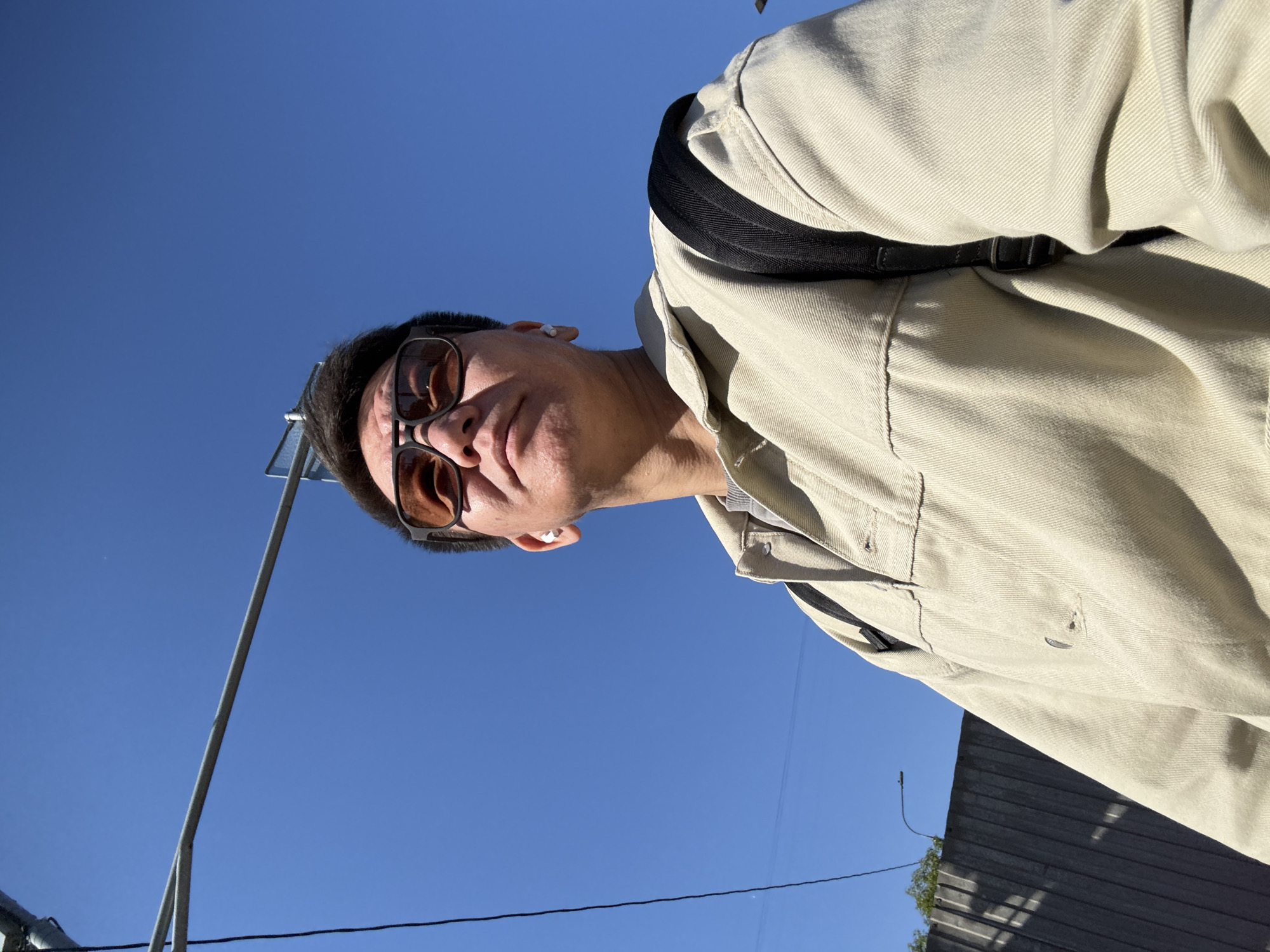 The image presents a low-angle outdoor shot of a person from the chest up, set against a vast, clear blue sky. The individual, appearing to be male, is looking slightly downwards and to the left of the frame (if rotated upright). He wears dark-framed sunglasses with brown-tinted lenses, obscuring his eyes. His mouth is closed with a neutral expression, and a white earbud is visible in his right ear.
He is dressed in a light-colored, possibly beige or pale yellow, long-sleeved button-up jacket made of a sturdy fabric. A black strap from a backpack is visible over his right shoulder. Bright sunlight illuminates his face and jacket, casting shadows that emphasize the textures of his clothing and indicate a sunny day.
The background is dominated by a vibrant, cloudless blue sky. In the lower left, a thin, light-colored metallic pole or structure extends upwards. A thin black wire runs horizontally across the bottom section of the image. To the lower right, a dark, corrugated surface, resembling a fence or building wall, is partially visible, with a hint of green foliage beneath it. The overall perspective is from below, looking up at the subject. There is no visible text in the image.
