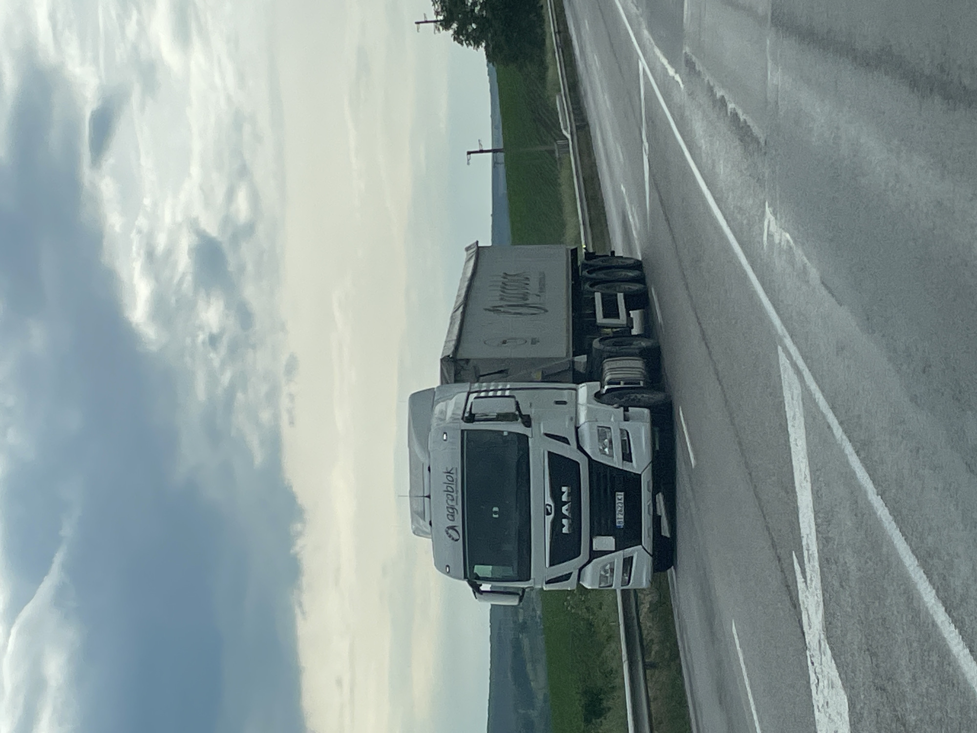 The image depicts a white MAN truck with an attached trailer on a highway near Beli Lom, Bulgaria. The truck has the "agroblok" logo and the license plate "BT 7620 KI." The trailer has a logo on the side. The background shows a cloudy sky, green fields, and power lines. The highway has road markings.