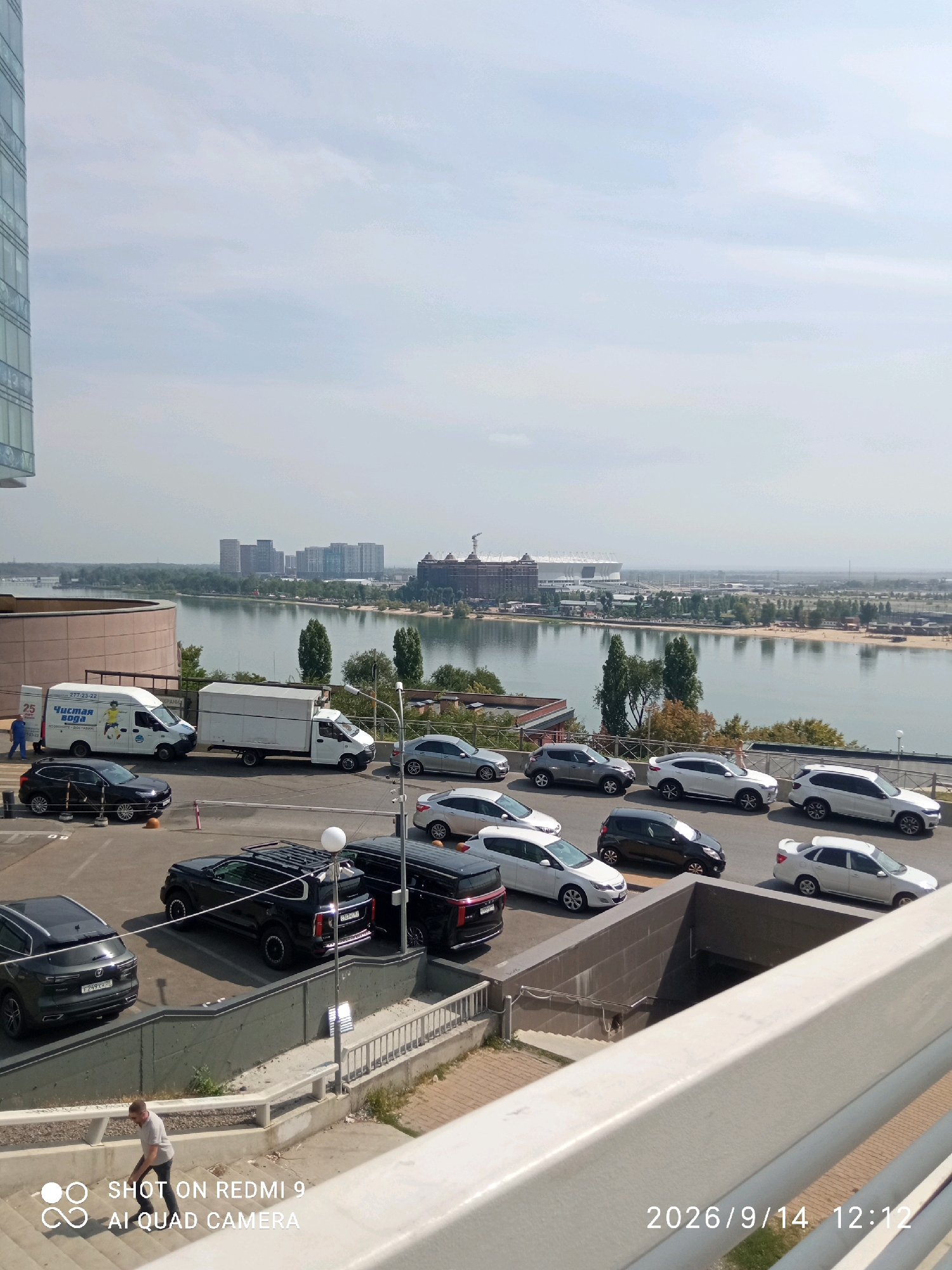 This image shows a view of the Don River in Rostov-on-Don, Russia, with a modern stadium visible in the distance. In the foreground, there are several parked cars and a street with a person walking down stairs. The scene appears to be a city street with commercial buildings and a body of water. The lighting suggests it is daytime, with a slightly overcast sky.