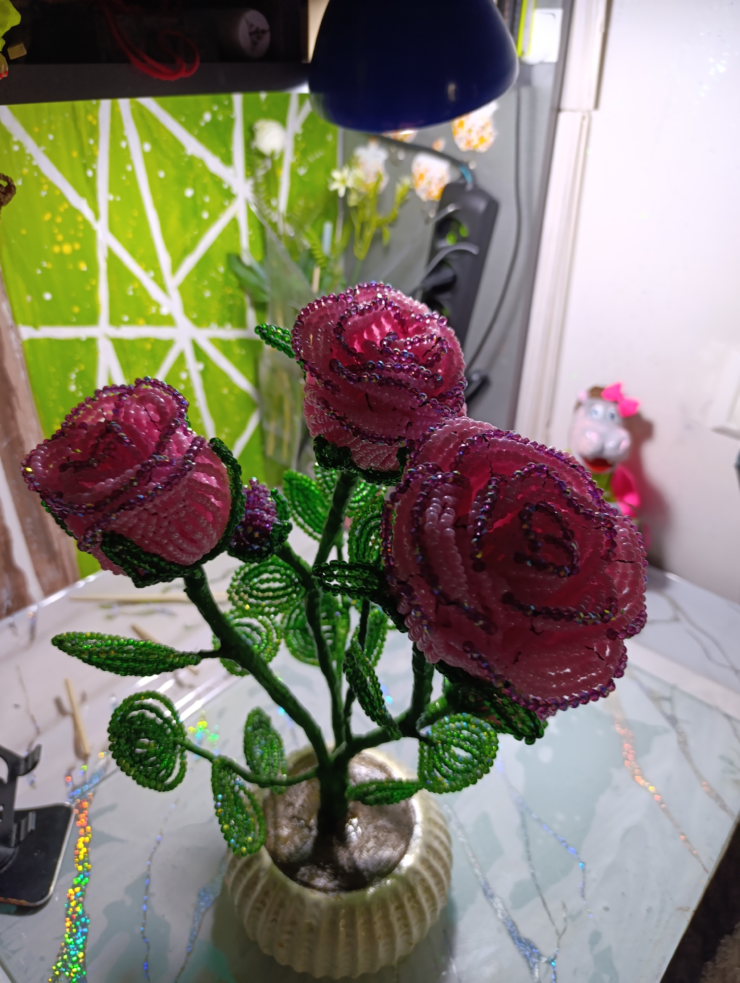 This media file features a close-up view of a beaded craft depicting three pink roses with green leaves and stems, planted in a decorative pot. The roses appear to be made from small pink and purple beads, with iridescent highlights. The leaves and stems are crafted from green beads. The arrangement is placed on a table with a light-colored, patterned surface that reflects light.
In the background, a vibrant green canvas with white abstract lines and yellow speckles is visible. To the right, a blurred image of a toy, possibly a hippopotamus with a pink bow, is present. A blue lamp is also visible in the upper portion of the image, casting light. The overall setting appears to be an indoor space, likely a crafting or display area, with the objects positioned on a surface that suggests a home or studio environment. There is no indication of time of day or weather. No visible text is present in the image. The location context provided is Fergana City, Uzbekistan, but this cannot be visually confirmed from the image itself.